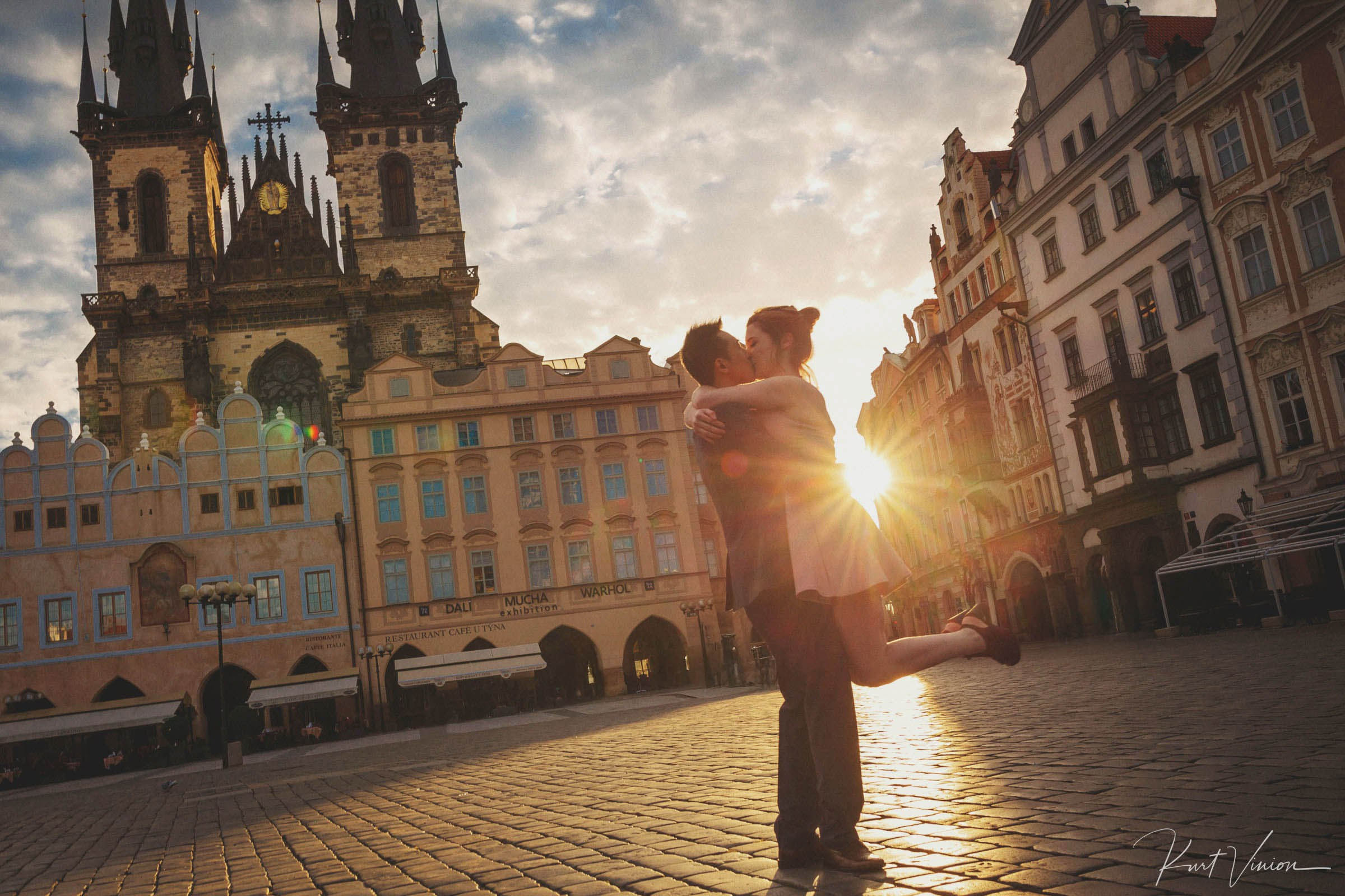 Man lifting fiancé giving kiss in golden sunlight in Prague Old Town Square.