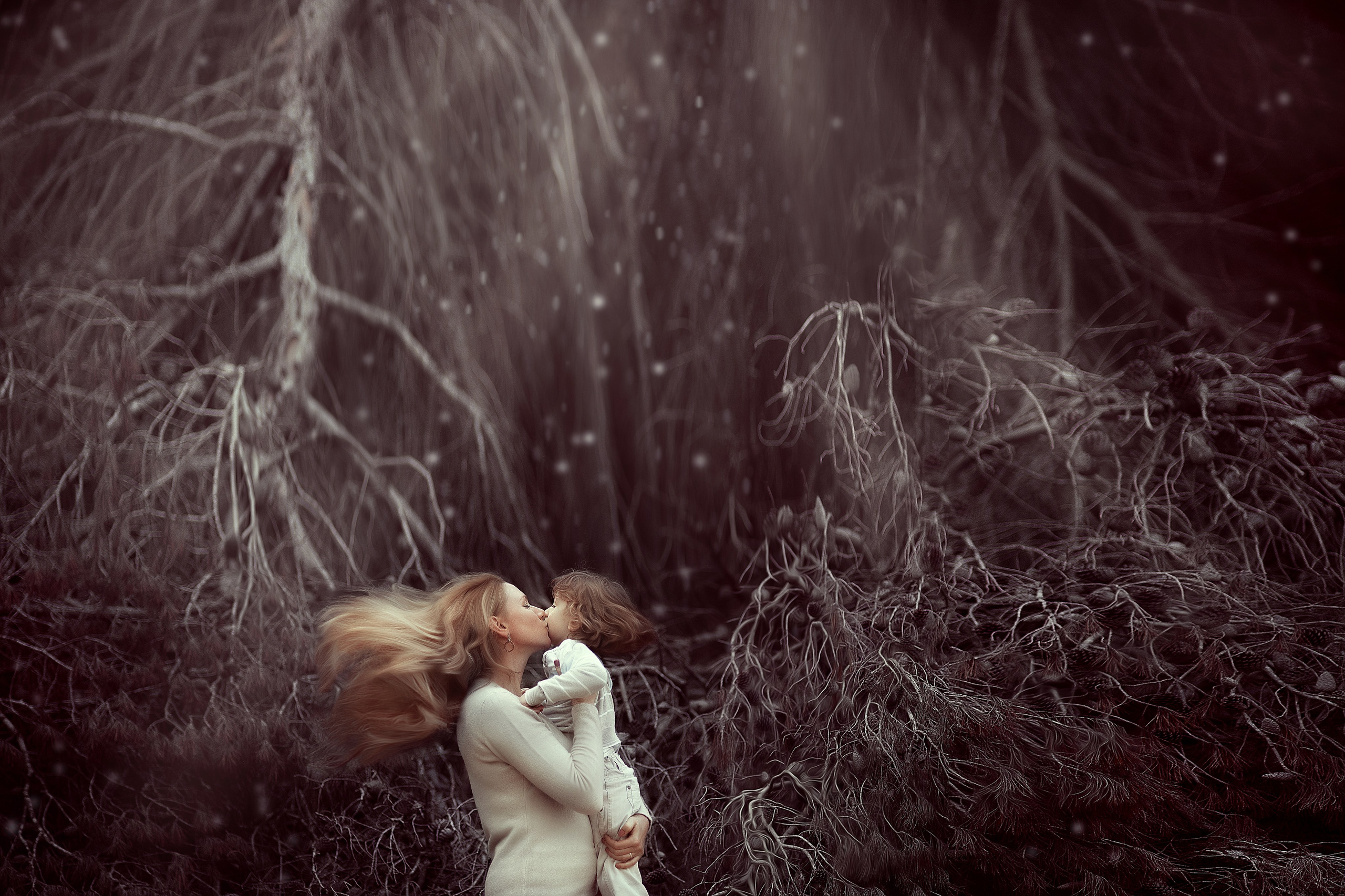 A blonde mother kisses her toddler under a dramatic pine forest backdrop, with her long hair flowing in the wind.