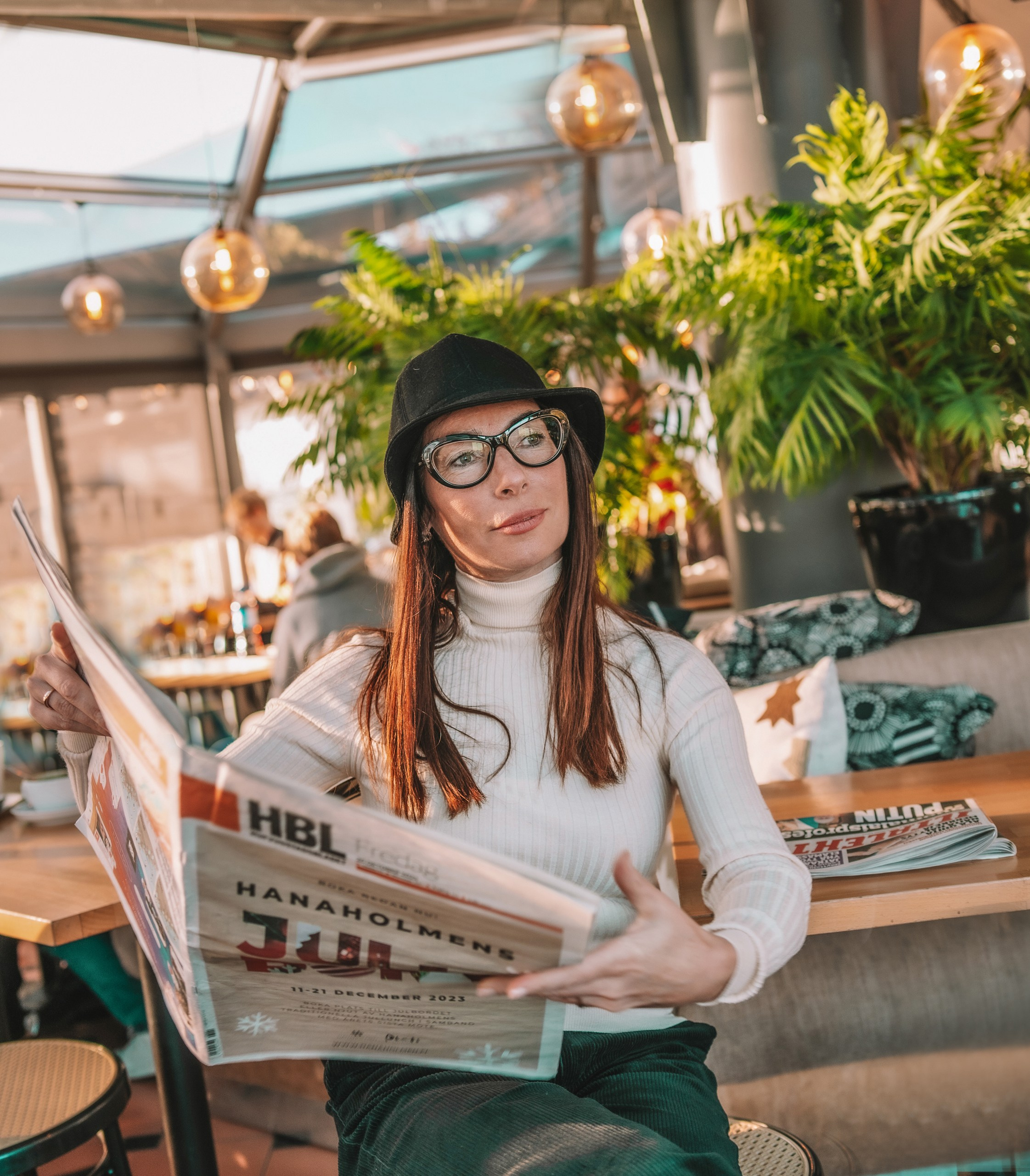 A woman in stylish black glasses and a hat reads a newspaper while sitting in a glass cafe, Riga portrait photographer