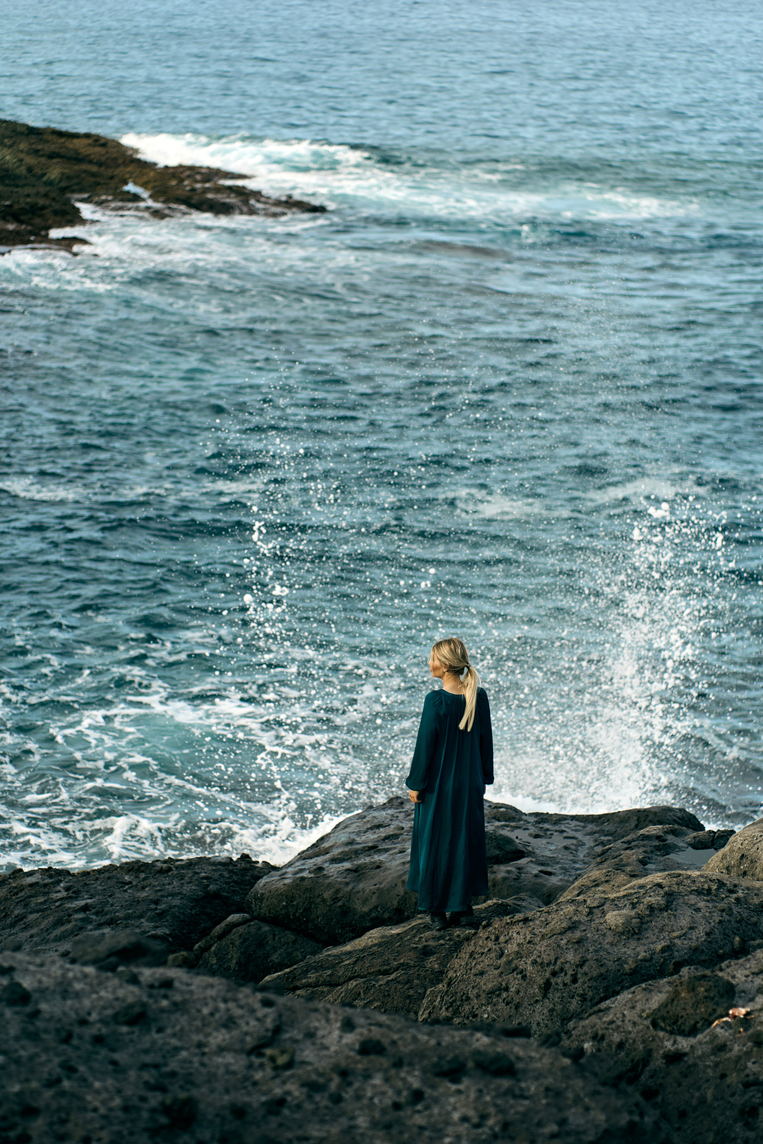 A solitary woman standing on rocky cliffs by the ocean, facing away, surrounded by the calm of nature.