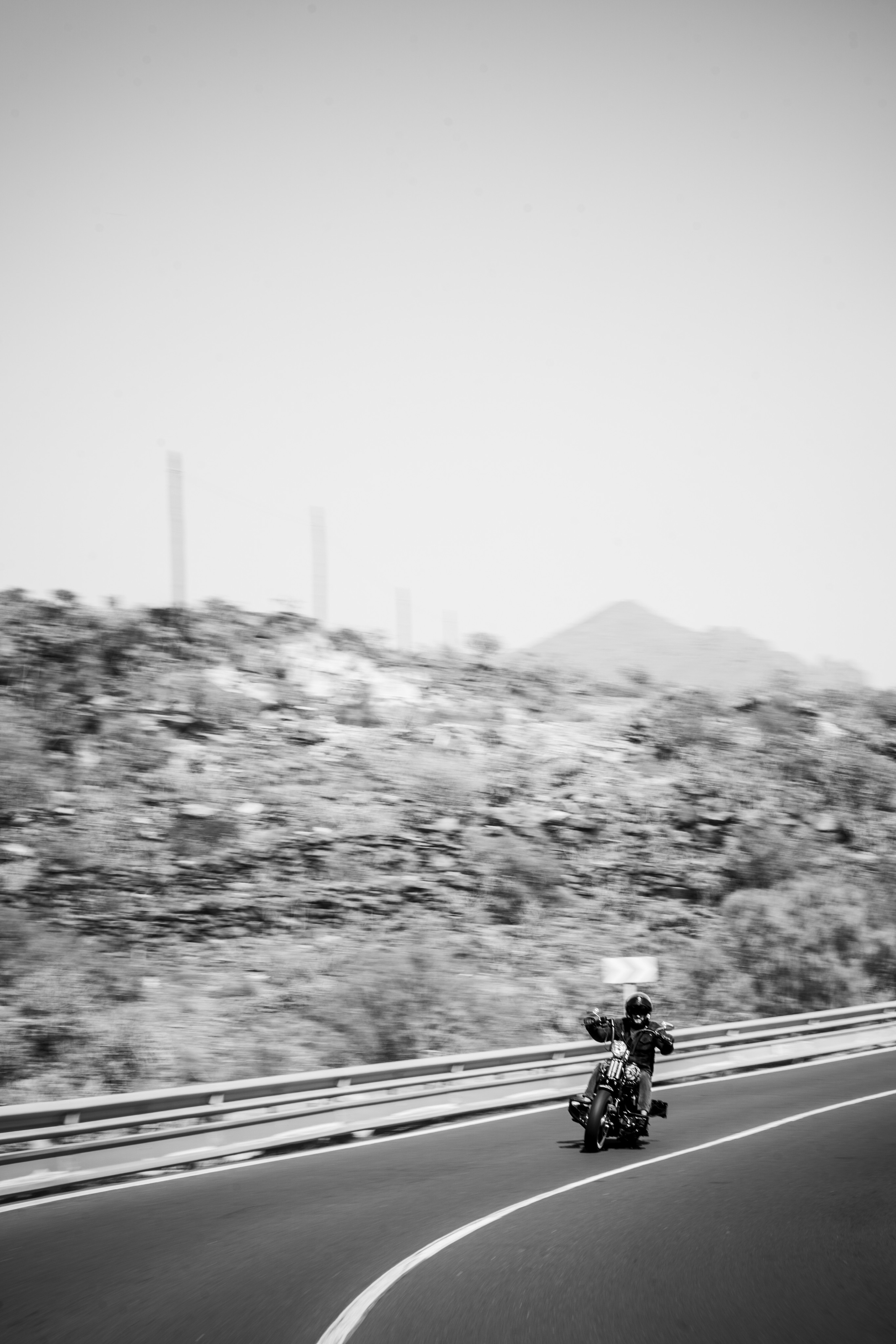 A black-and-white photo of a motorcyclist riding on a winding road through a barren, mountainous landscape.