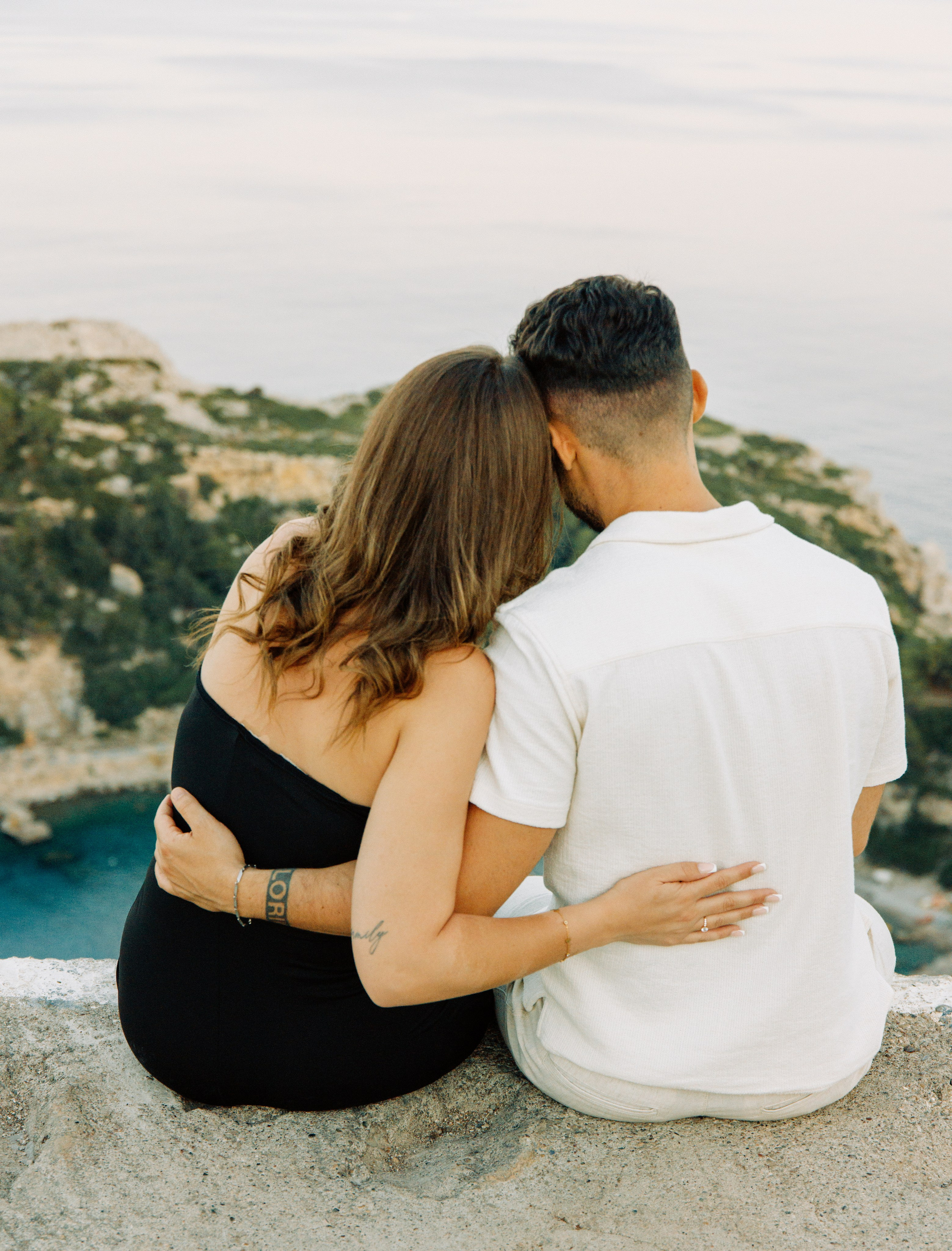 "Rückenansicht eines Paares, das nebeneinander auf den Felsen des Profitis Ilias sitzt. Sie schauen gemeinsam auf das tiefblaue Ägäische Meer und die Küste von Rhodos unter ihnen. Ein Moment des Innehaltens nach dem Heiratsantrag in der friedlichen Natur Griechenlands."