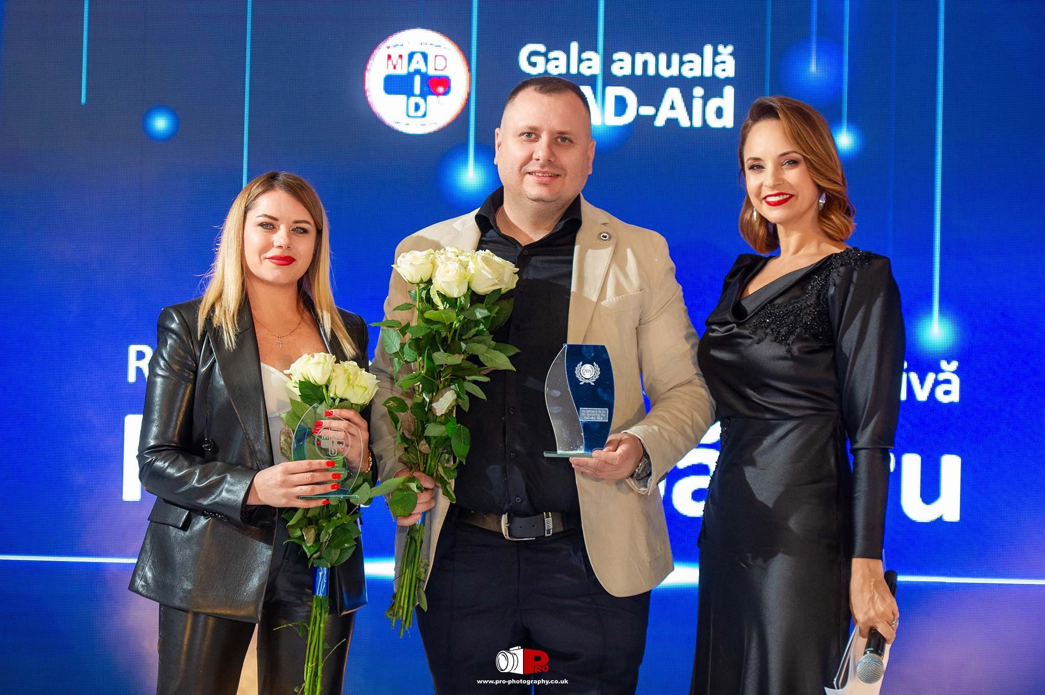 A man and two women proudly posing with awards and roses on stage at the MAD-Aid gala.