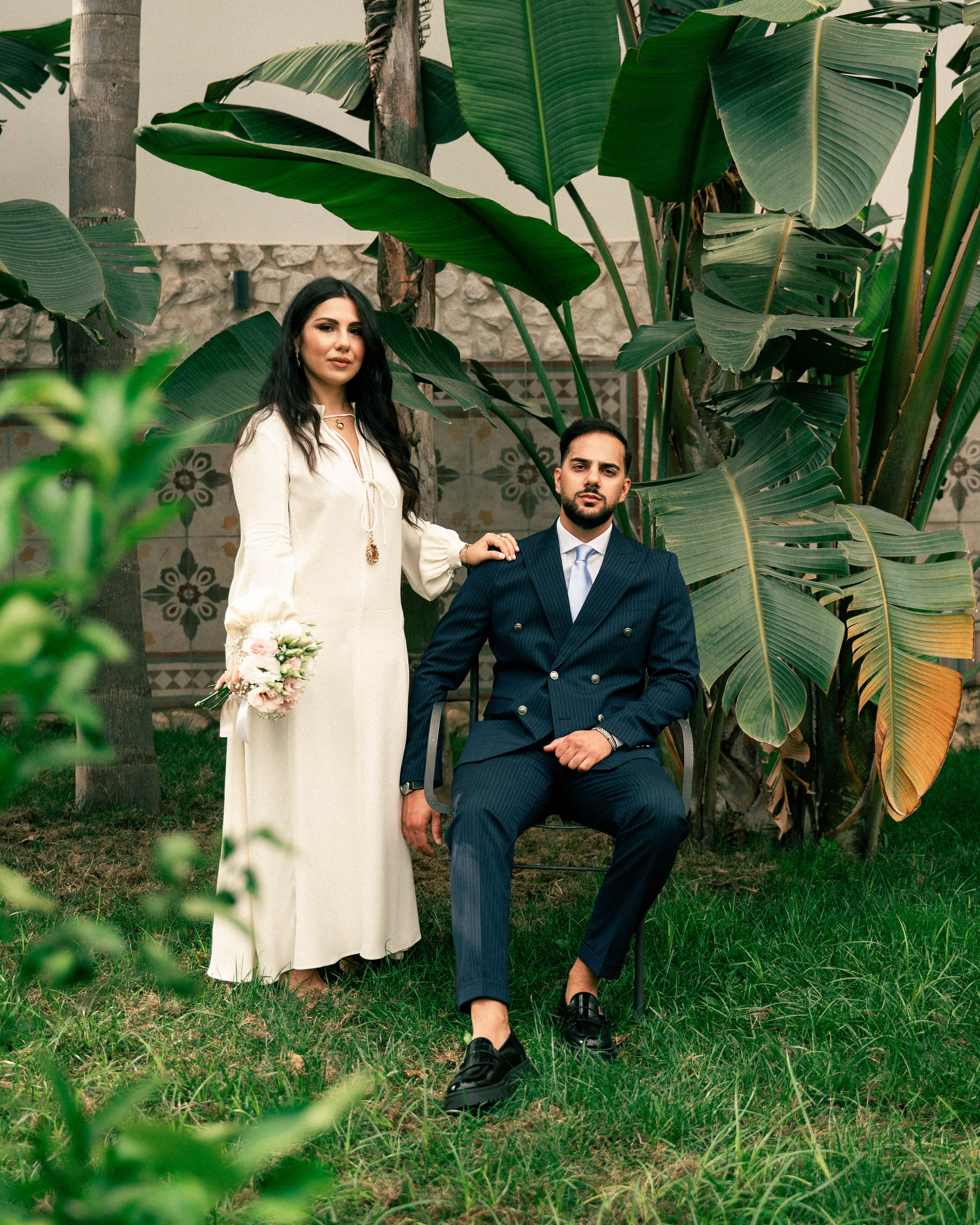 Bride standing next to groom seated on a chair in a garden setting