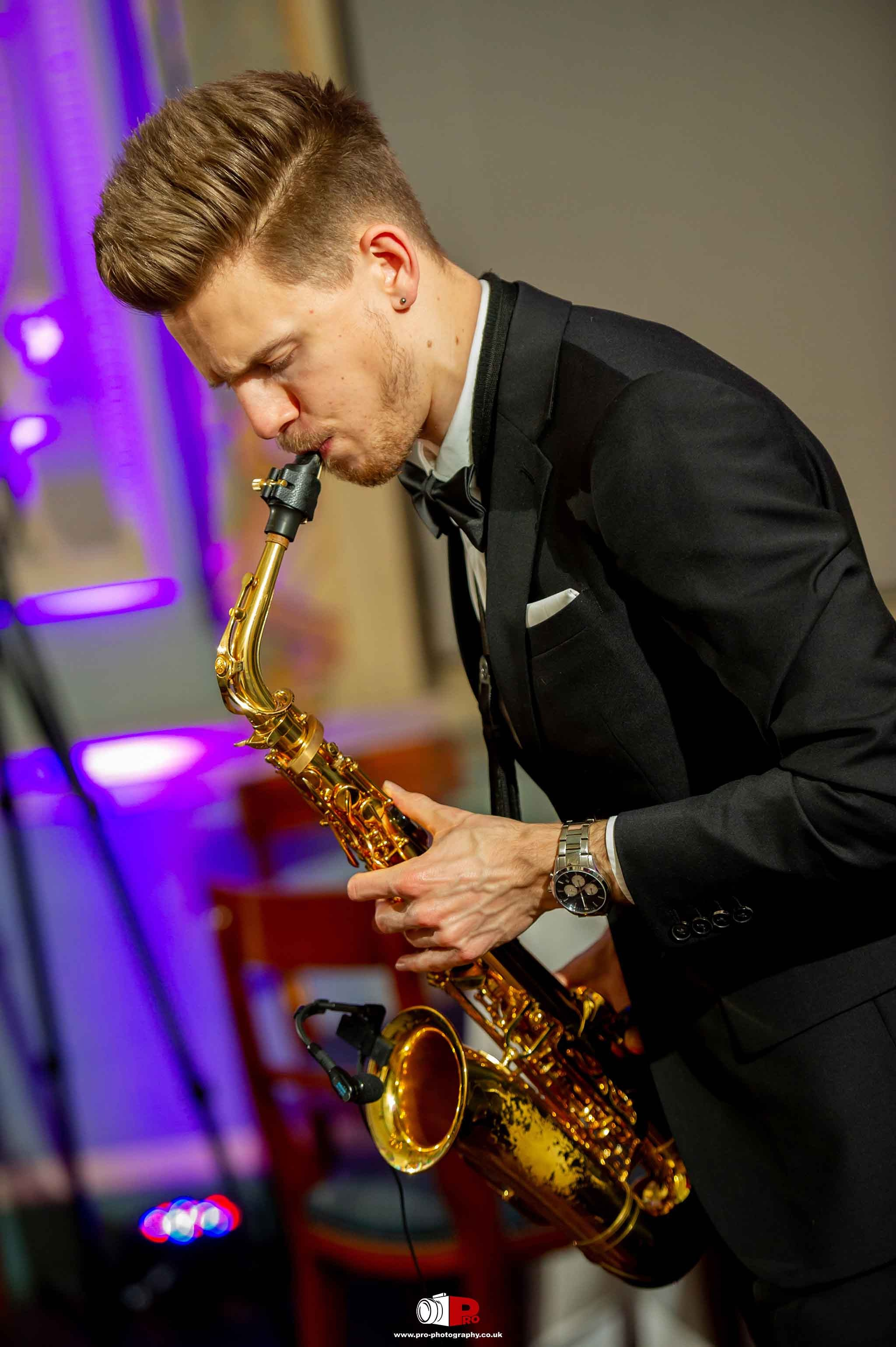 A young saxophone player in a black suit is performing on stage with vibrant purple lighting.