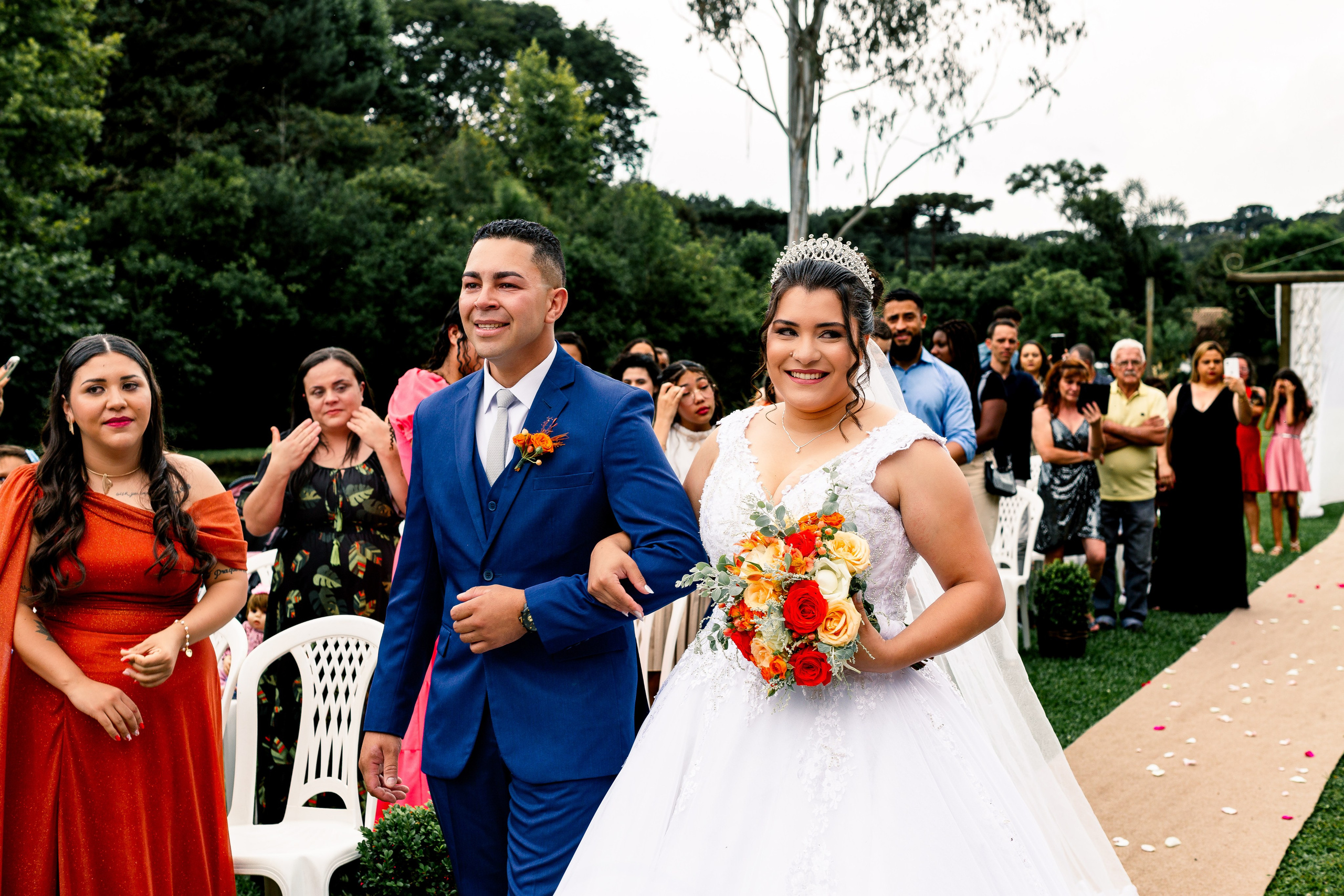 Recém-casados caminhando sorridentes pelo corredor sob aplausos, com convidados celebrando ao fundo.