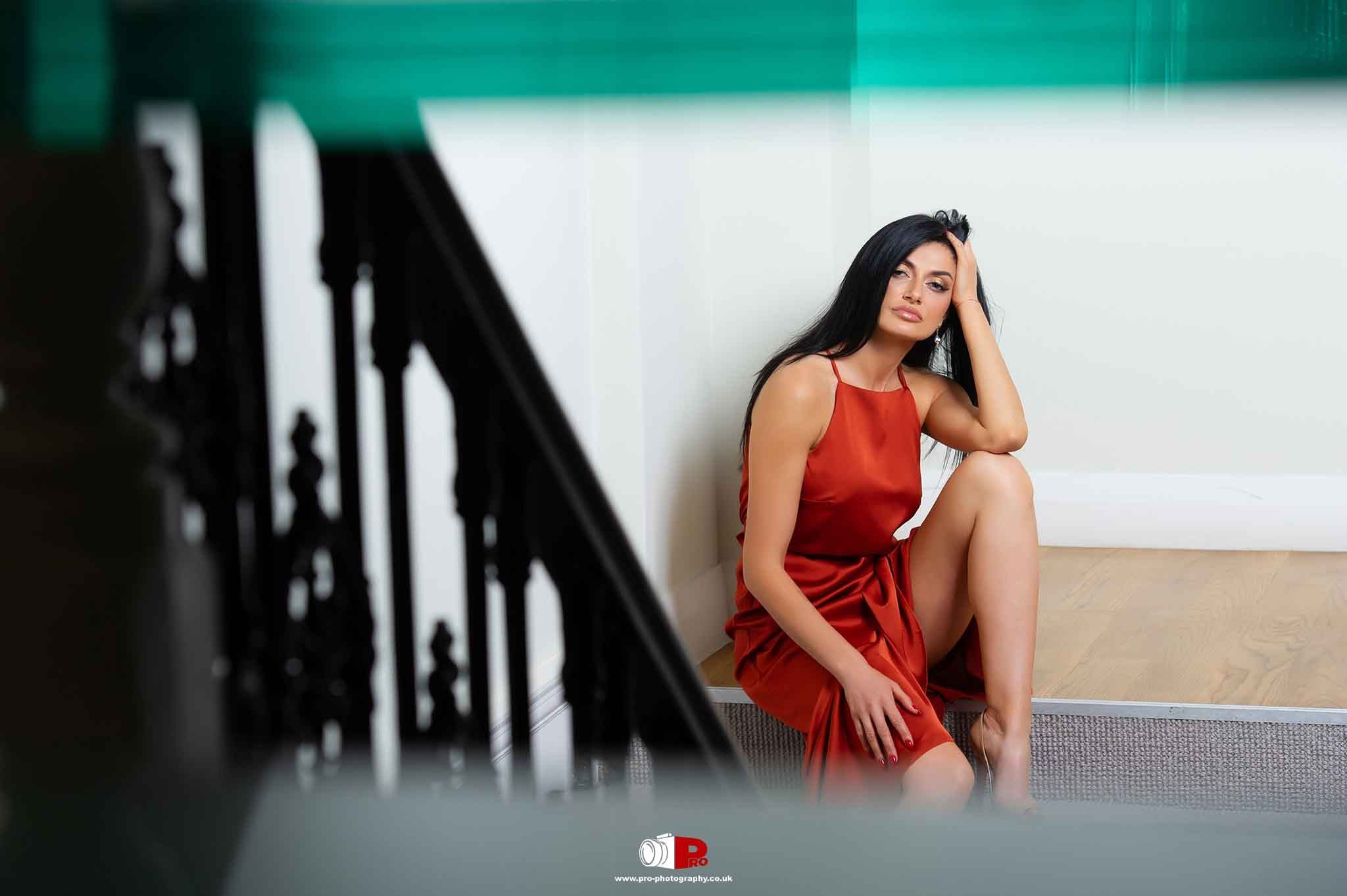A woman in a red dress sits thoughtfully on a staircase, gazing into the distance.