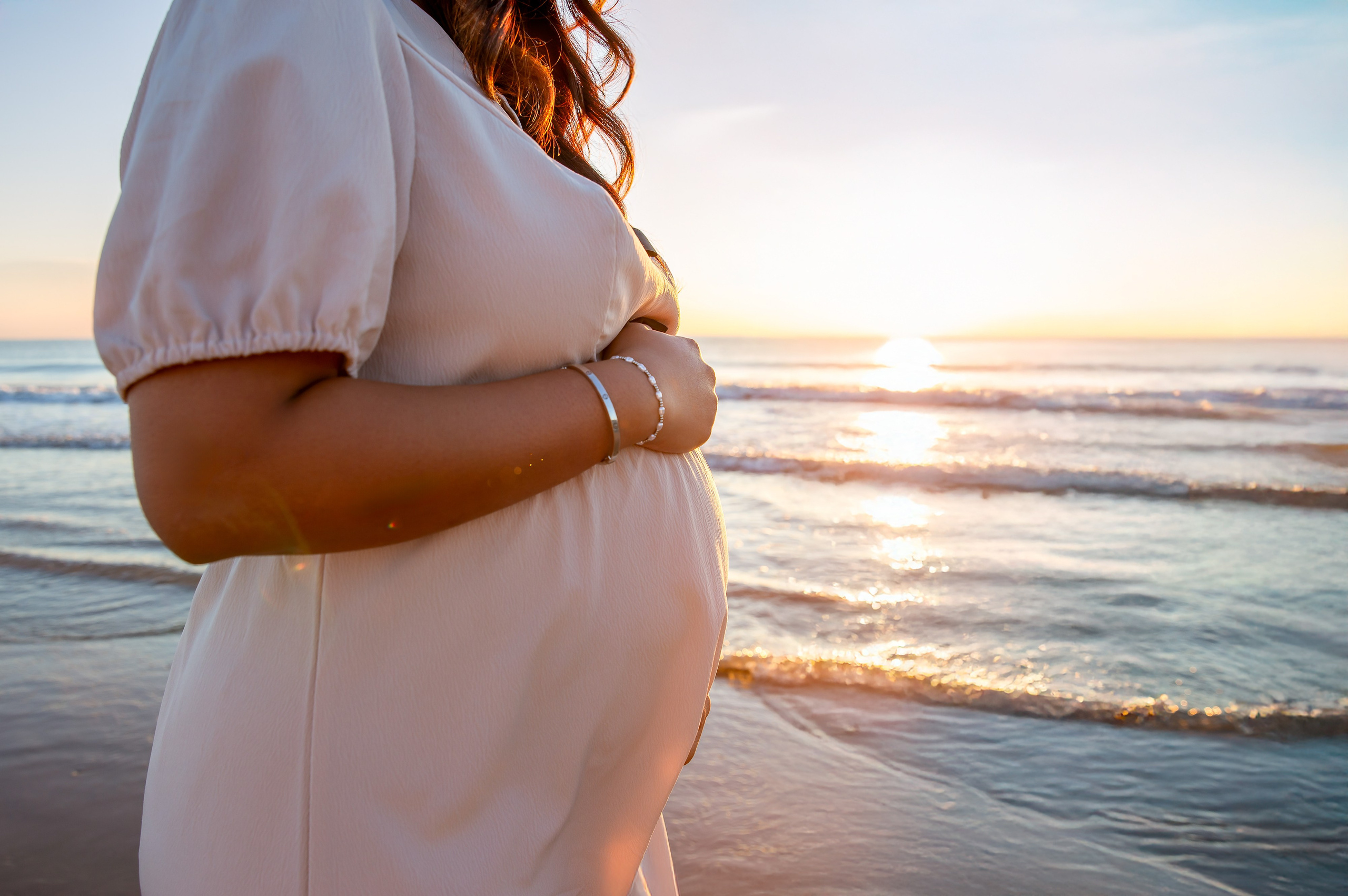 Primer plano de una mujer embarazada sosteniendo su barriga durante una sesión de fotos al atardecer en la playa de Valencia, España — ideal para quienes buscan sesiones de maternidad elegantes y emocionales en Valencia y en toda España.