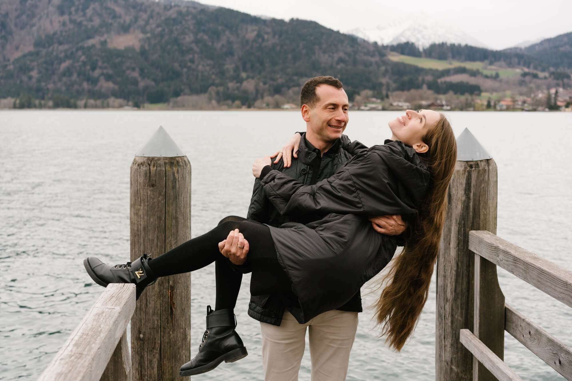 Couple standing side by side on a wooden dock at Tegernsee lake with mountains and bare trees during couple session