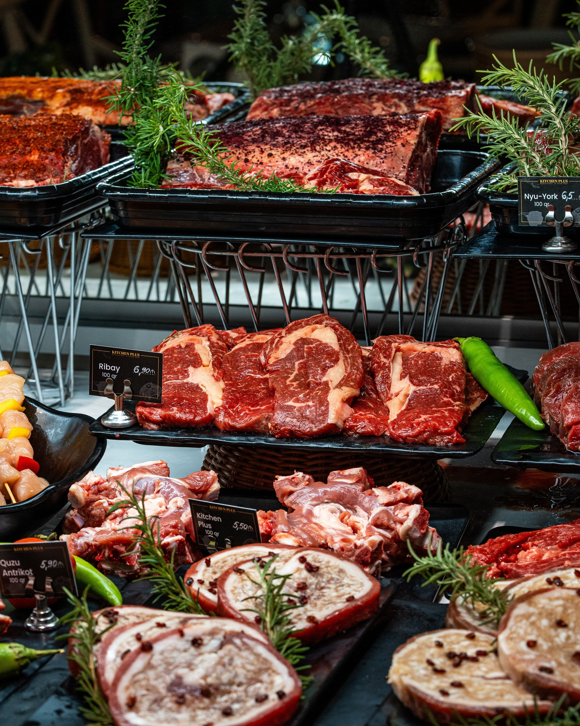 Qəssab vitrininin yaxın plan görünüşü, qiymət etiketləri ilə Ribeye və New York steykləri, quzu antrikotu (quzu kəsimləri), və döş əti rulonları.