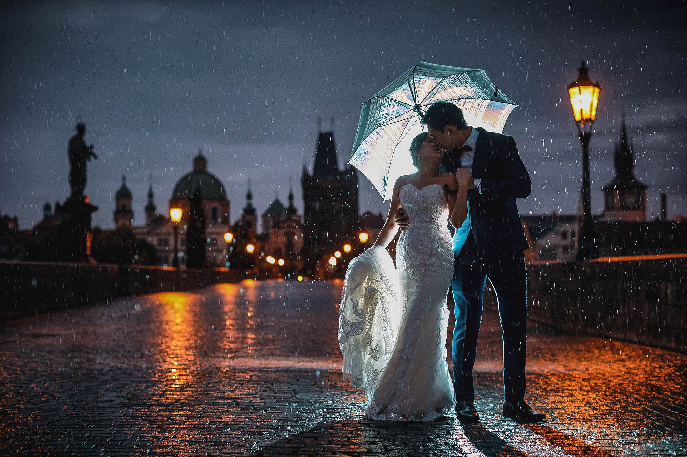 Elegantly dressed newlyweds embrace under an umbrella in the pouring rain atop the nighttime landscape of the Charles Bridge