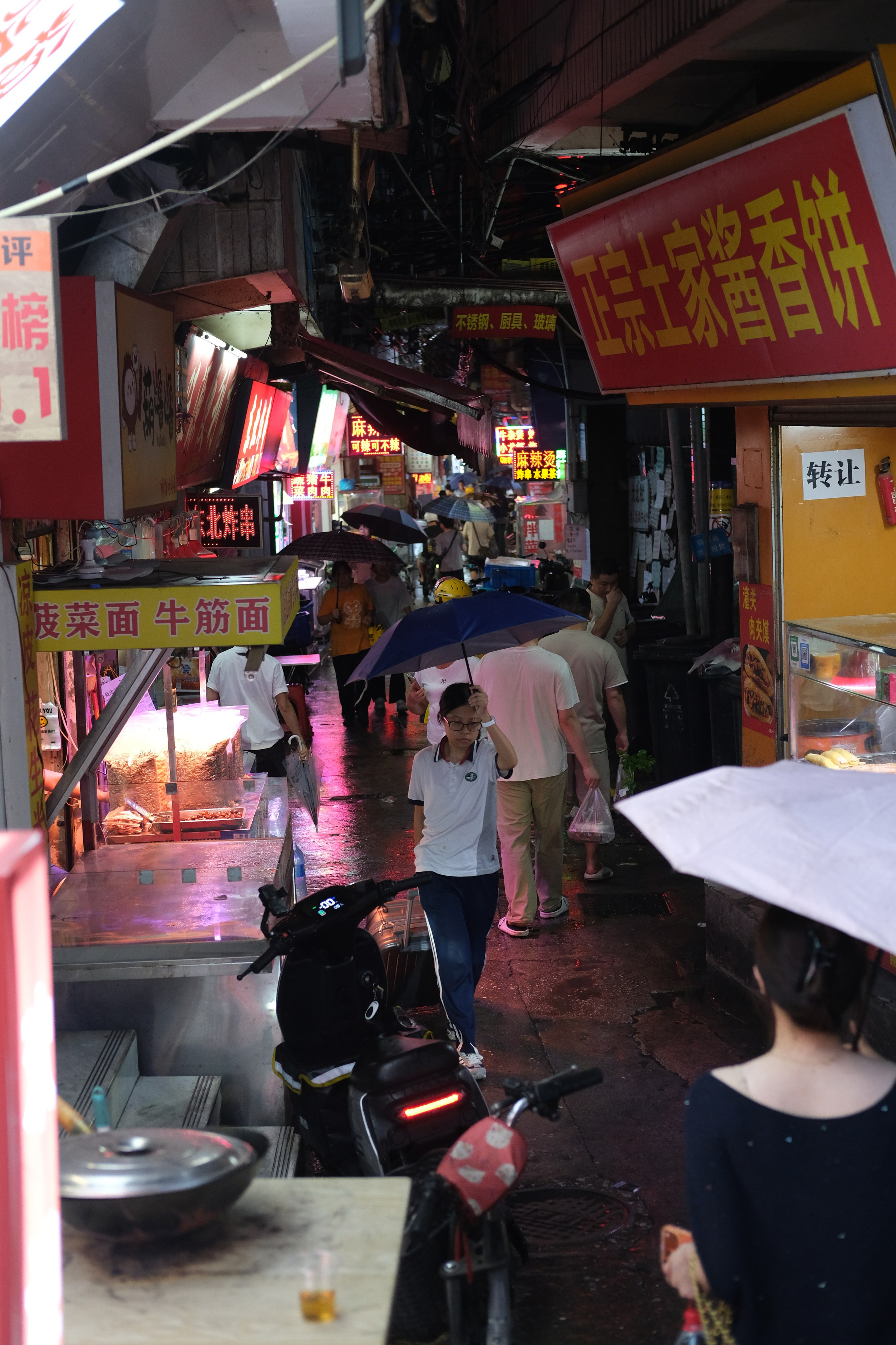 Rainy evening street scene in Guangzhou with pedestrians carrying umbrellas through a neon-lit market alley, photographed with a Fujifilm X-Pro3 and finished with a cinematic film-inspired edit.