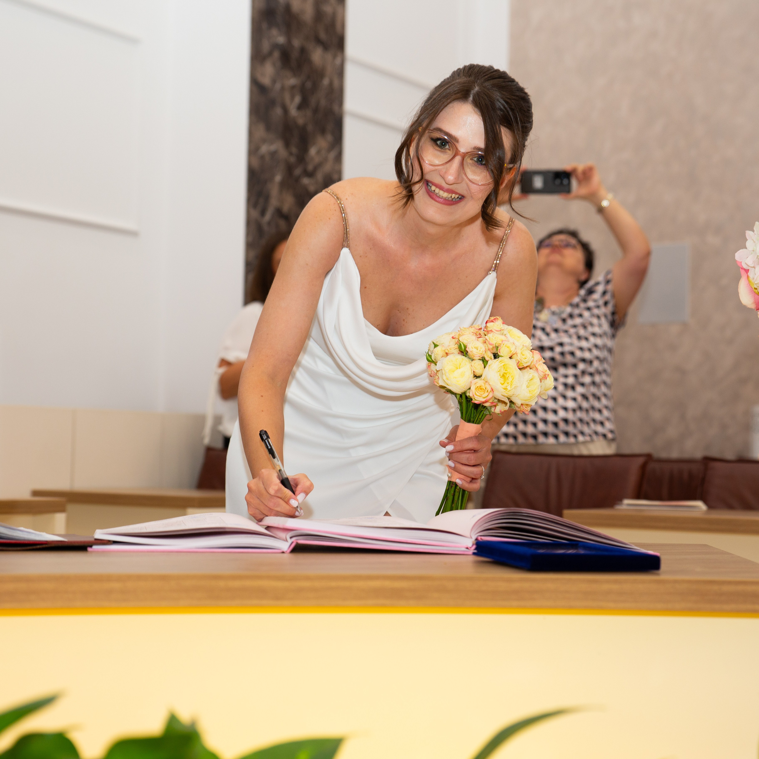 Bride in white dress with bouquet signing a register during a civil wedding ceremony.