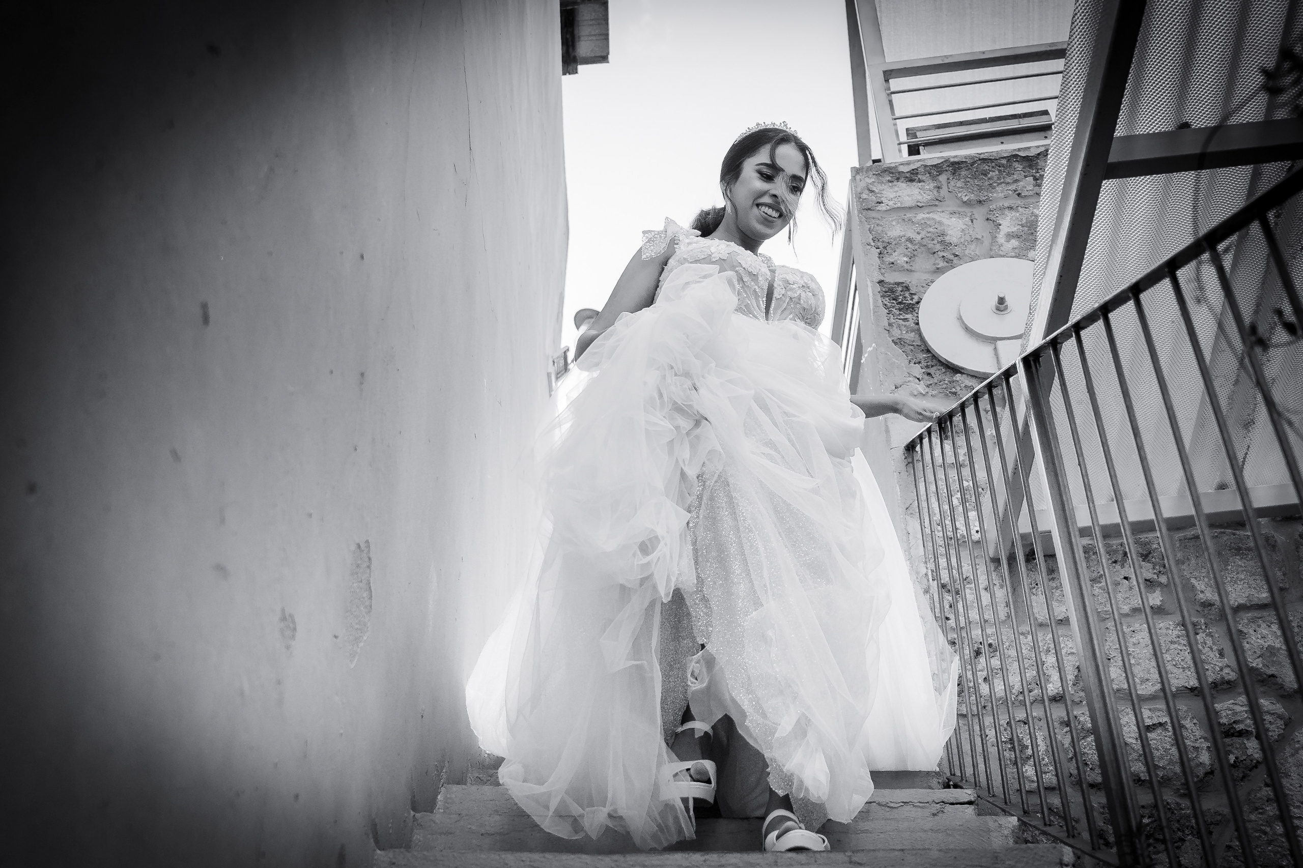 A black-and-white photo of the bride descending a stone staircase in a wedding dress with layered ruffles. She is smiling and looking down, with blurred architectural elements of a city or historical building in the background.