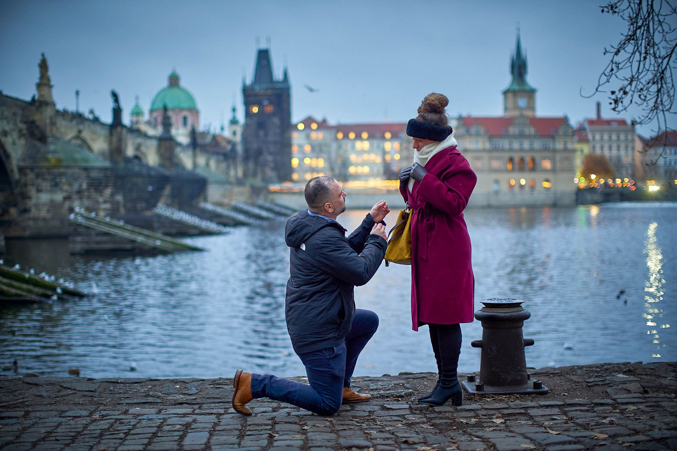 Man on knee holding ring to woman as she stands in disbelief against the backdrop of the Charles Bridge.