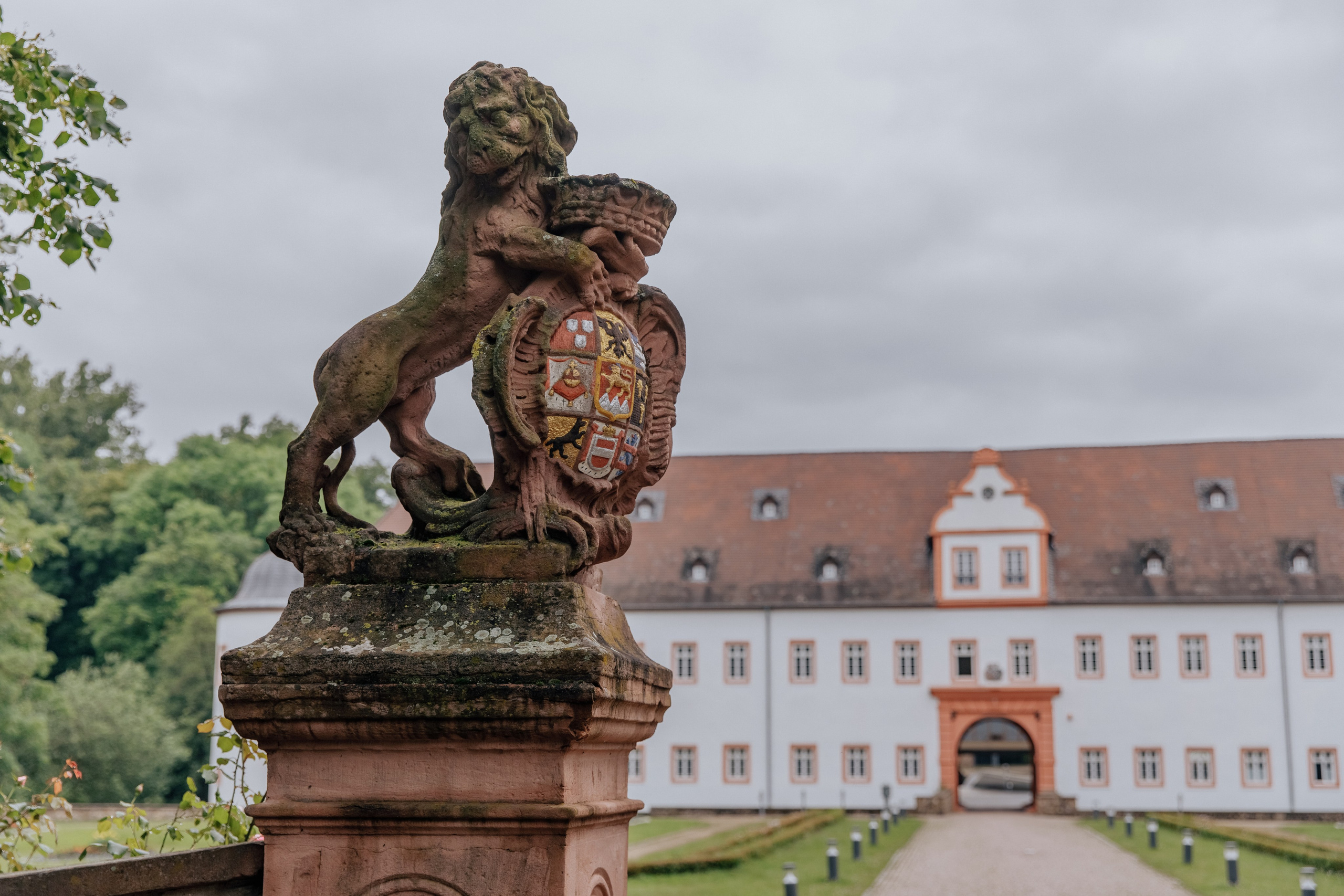 Statue eines Löwen mit einem Schild im Vordergrund, mit dem Schloss Heusenstamm im Hintergrund.