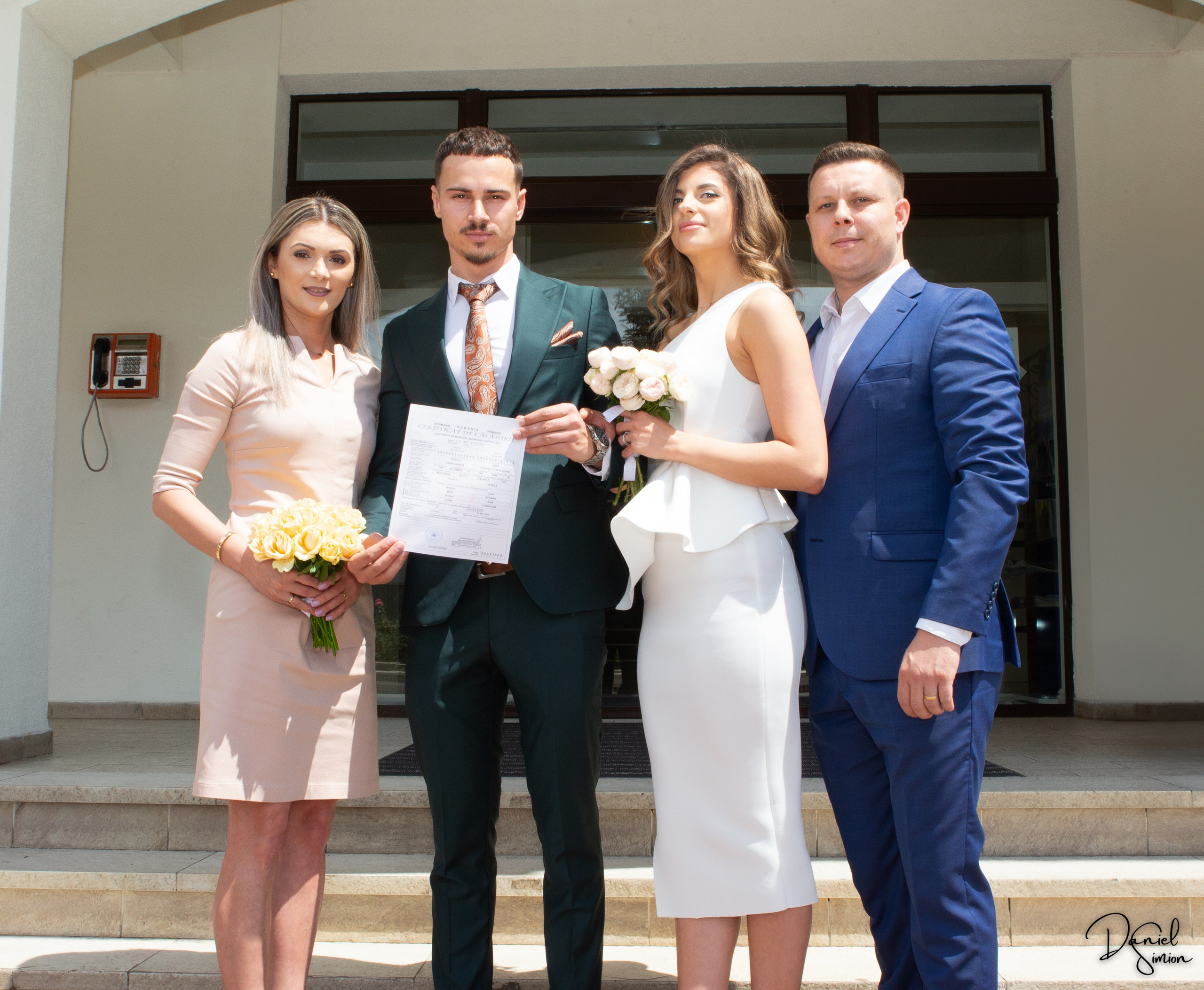 Newlywed couple and two witnesses standing outdoors holding a marriage certificate and bouquets.
