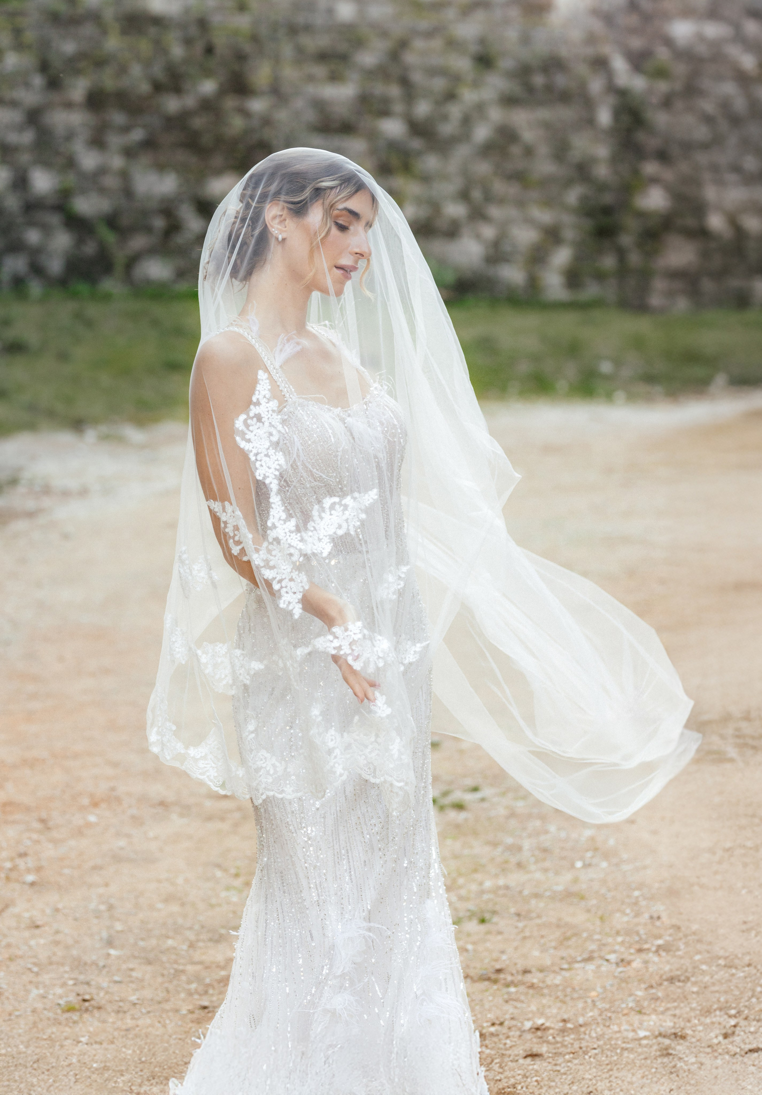 A radiant bride stands in the narrow, sunlit streets of Rhodes' Old Town, her intricate lace wedding dress contrasting beautifully with the weathered stone walls behind her. The editorial-style portrait highlights her serene expression and the timeless elegance of the historic setting, with soft natural light accentuating her features.