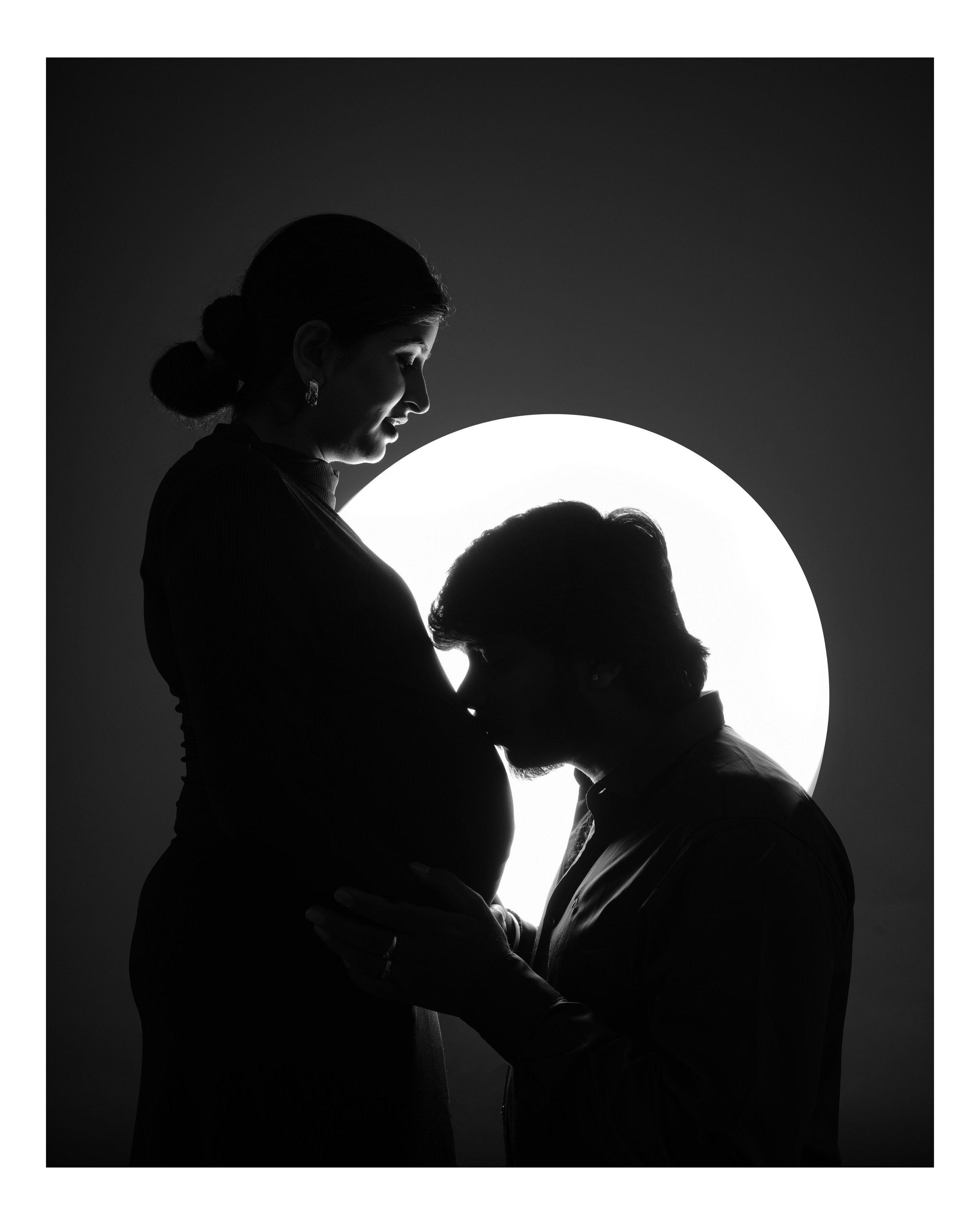 black and white studio silhouette portrait of a man kissing a pregnant woman's belly with a circular light in Malleshwaram, Bengaluru
