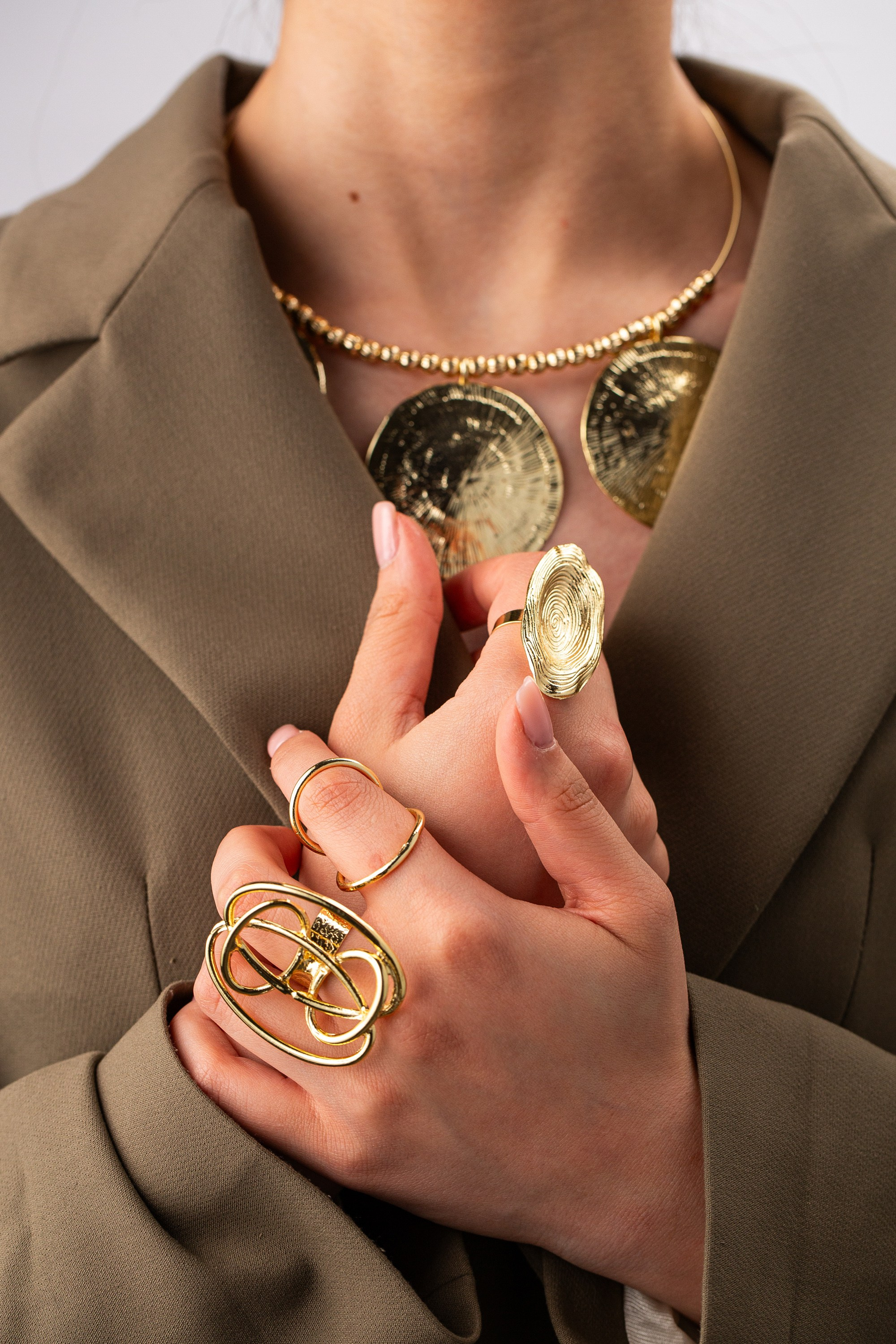 Cəsur qızıl zərgərlik ilə əllərin yaxından görünüşü. Üzüklər böyük və mücərrəddir və çəkiclə işlənmiş qızıl diskləri olan boyunbağı görünür.