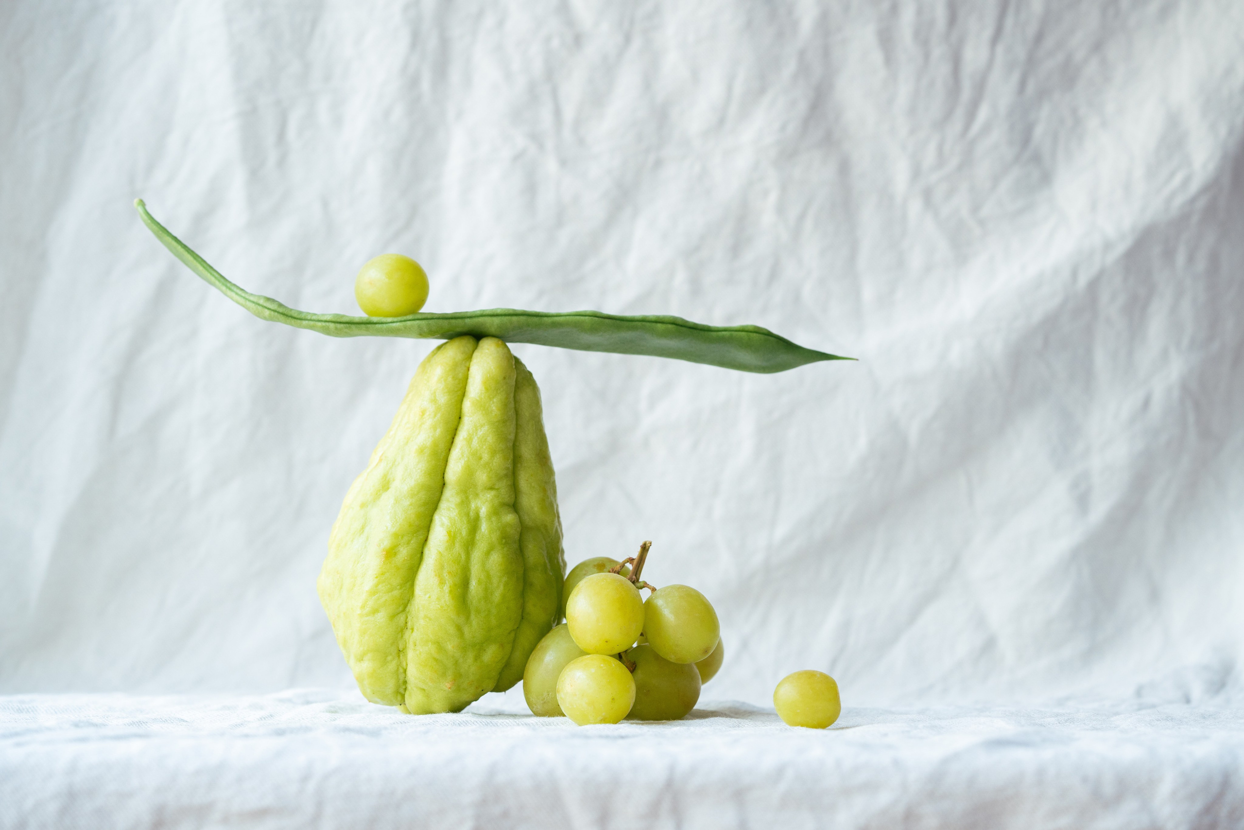 Balanced Still Life of Chayote, Green Beans, and Grapes. Artistic arrangement of fresh chayote, green beans, and grapes balanced on a white textured fabric background.