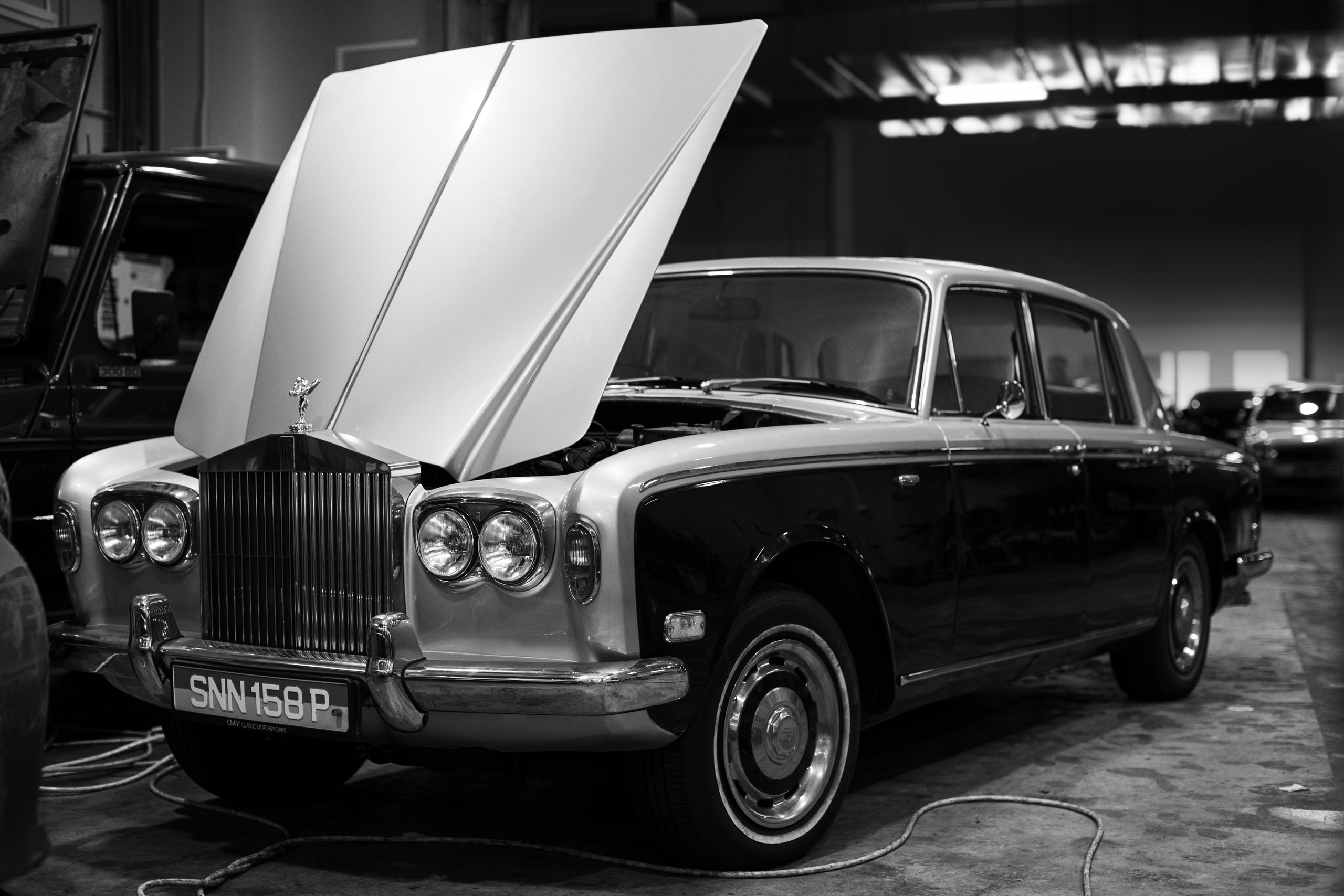 Black-and-white photograph of a classic Rolls-Royce with its hood raised, revealing intricate engine details at The Garage by Classic Motorworks in Singapore. Shot by Mirrorklezz Photography, this image captures the elegance and mechanical craftsmanship of the vintage Rolls-Royce, highlighting the iconic Spirit of Ecstasy hood ornament and the car's timeless design. Ideal for enthusiasts of classic car restoration and luxury automotive photography. https://www.classicmotorworks.com.sg/