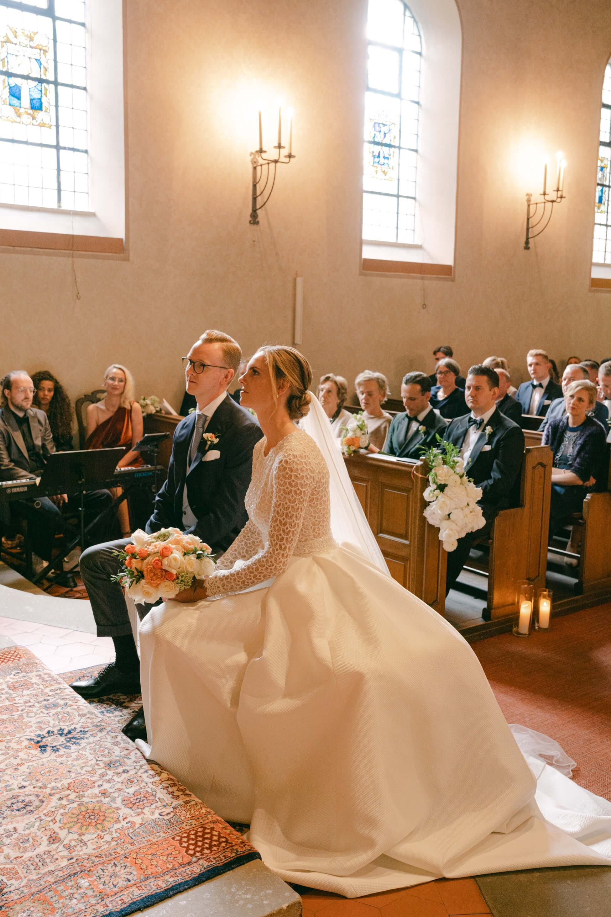 Bride in long-sleeve lace wedding dress seated at church ceremony holding bouquet, luxury wedding Frankfurt
