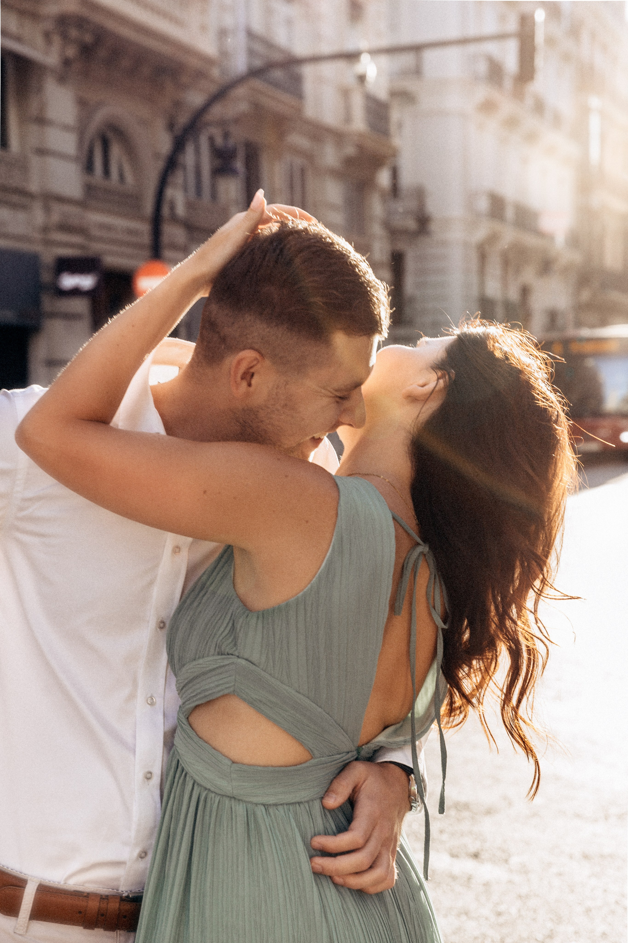 Romantic love story photo of a couple sharing an intimate kiss in the golden sunlight on a busy street in Valencia, Spain, with elegant historic architecture in the background.