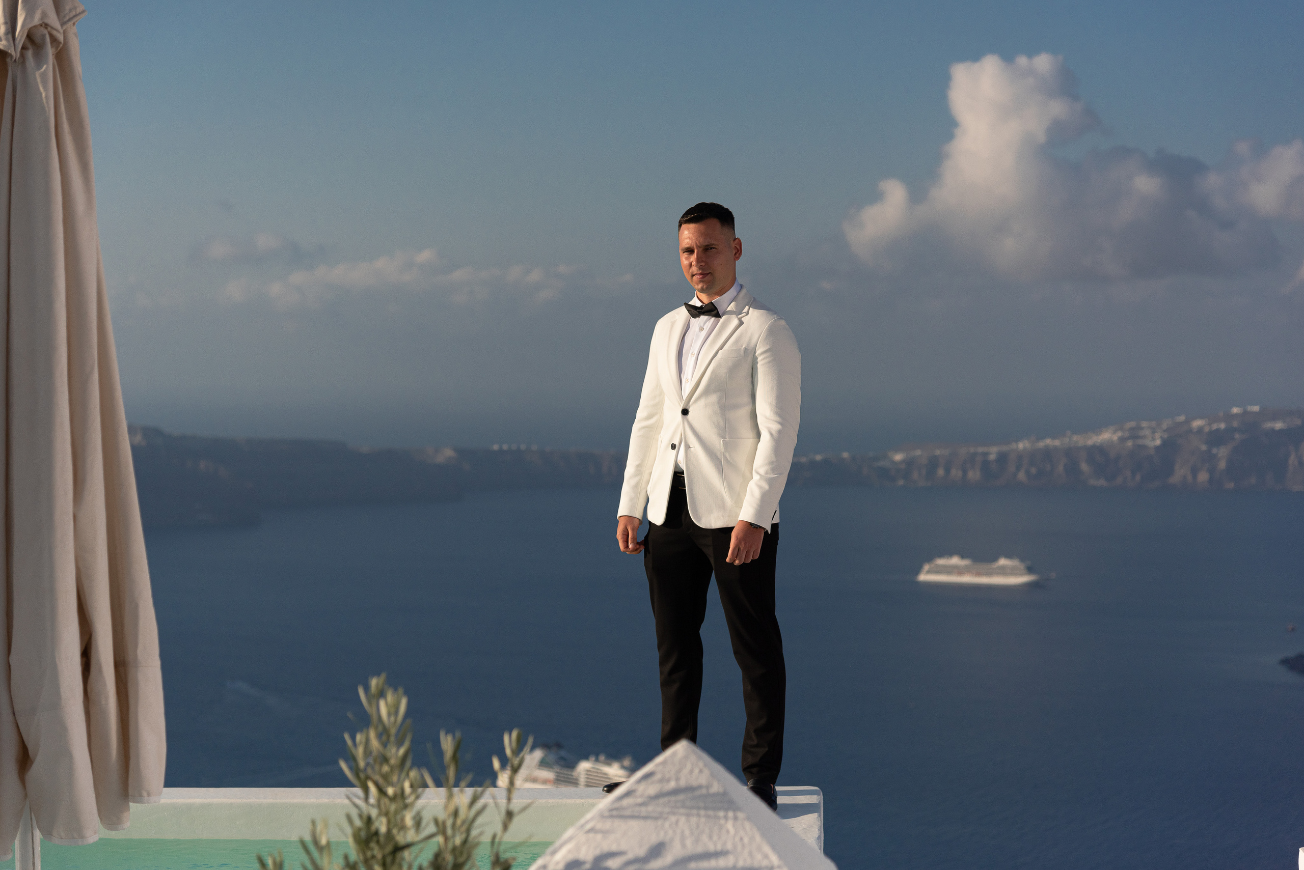 The groom in a white suit stands against the backdrop of the Santorini caldera