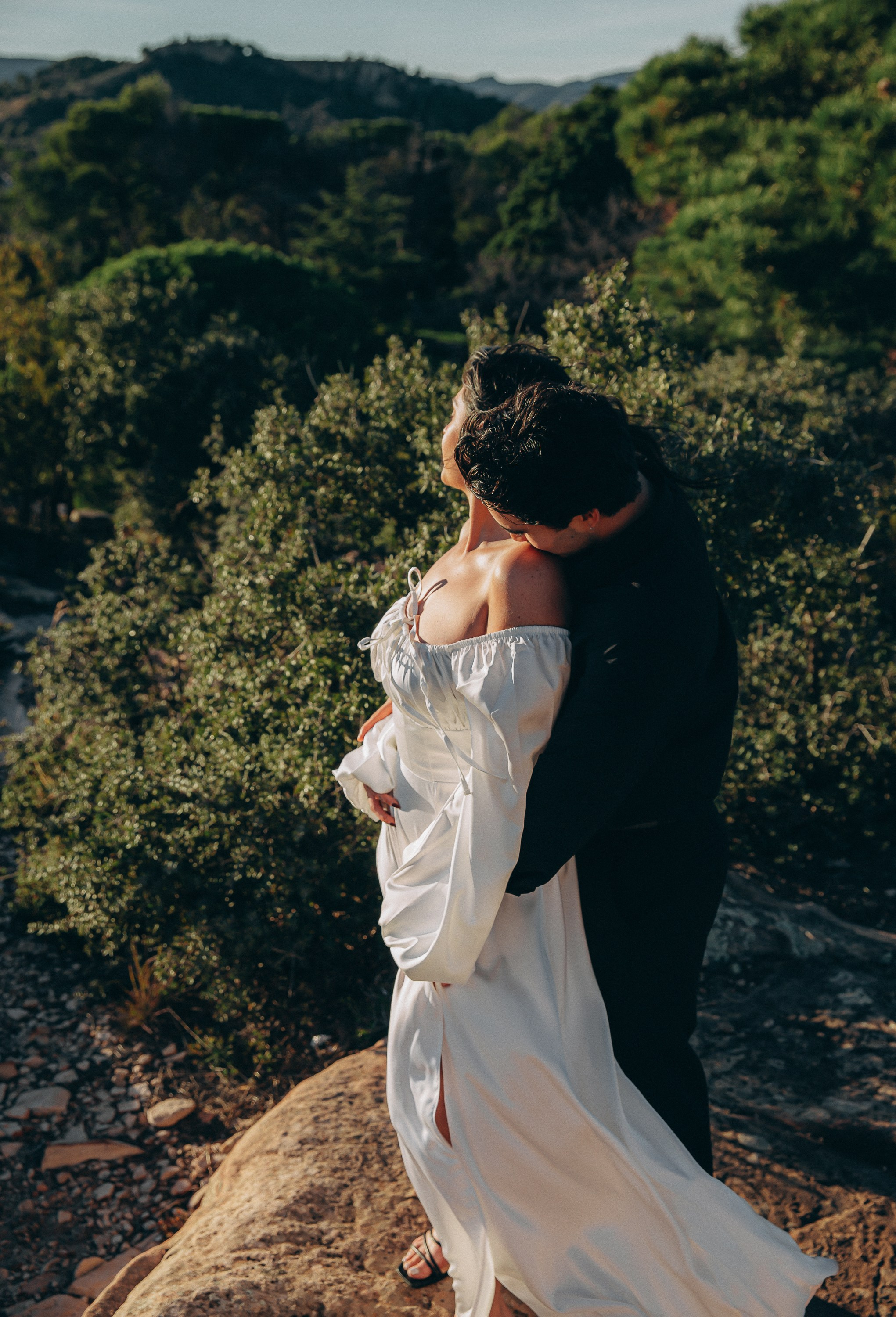 Couple hugging on a mountain cliff during a destination wedding in Barcelona, Spain, with lush green hills in the background. This intimate elopement portrait reflects the joy and connection of a romantic engagement turned wedding celebration.