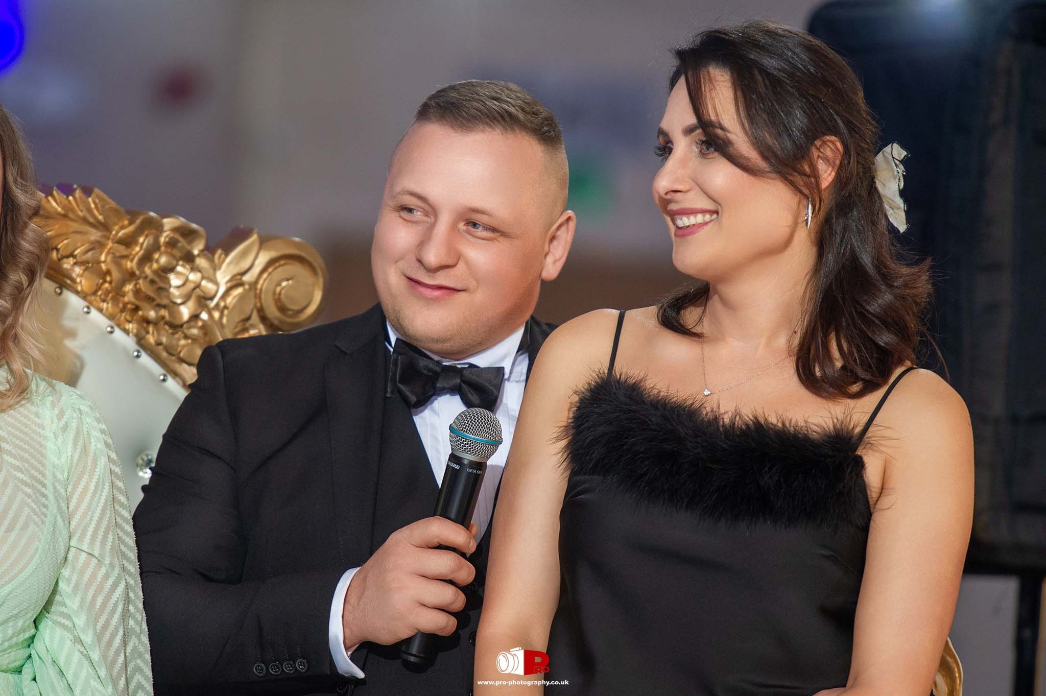 A smiling couple dressed formally is seated on a golden ornate chair at a prestigious gala event.