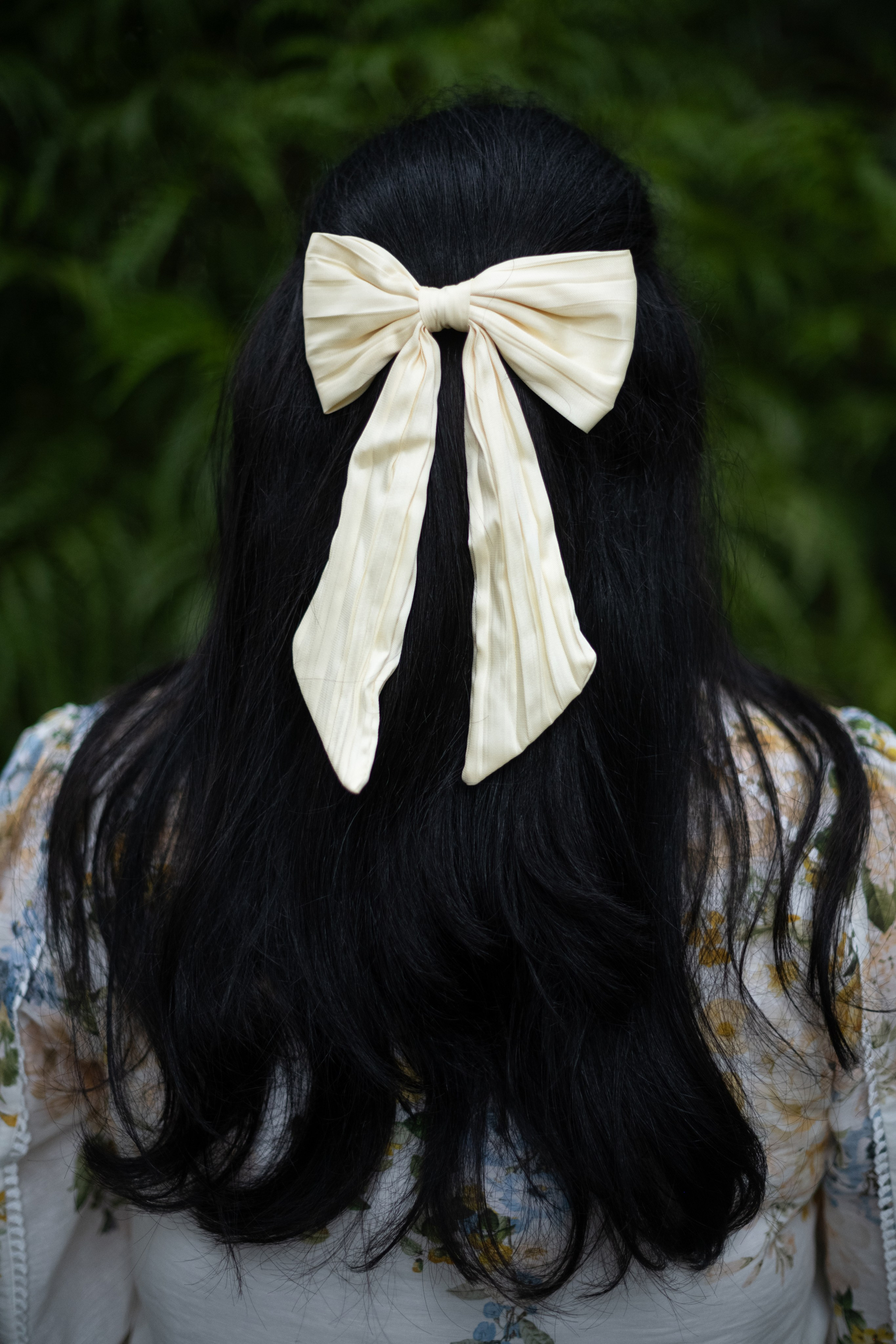 Rear view of long black hair adorned with an ivory bow, framed by fern foliage and soft floral-print sleeves during an outdoor maternity portrait session.