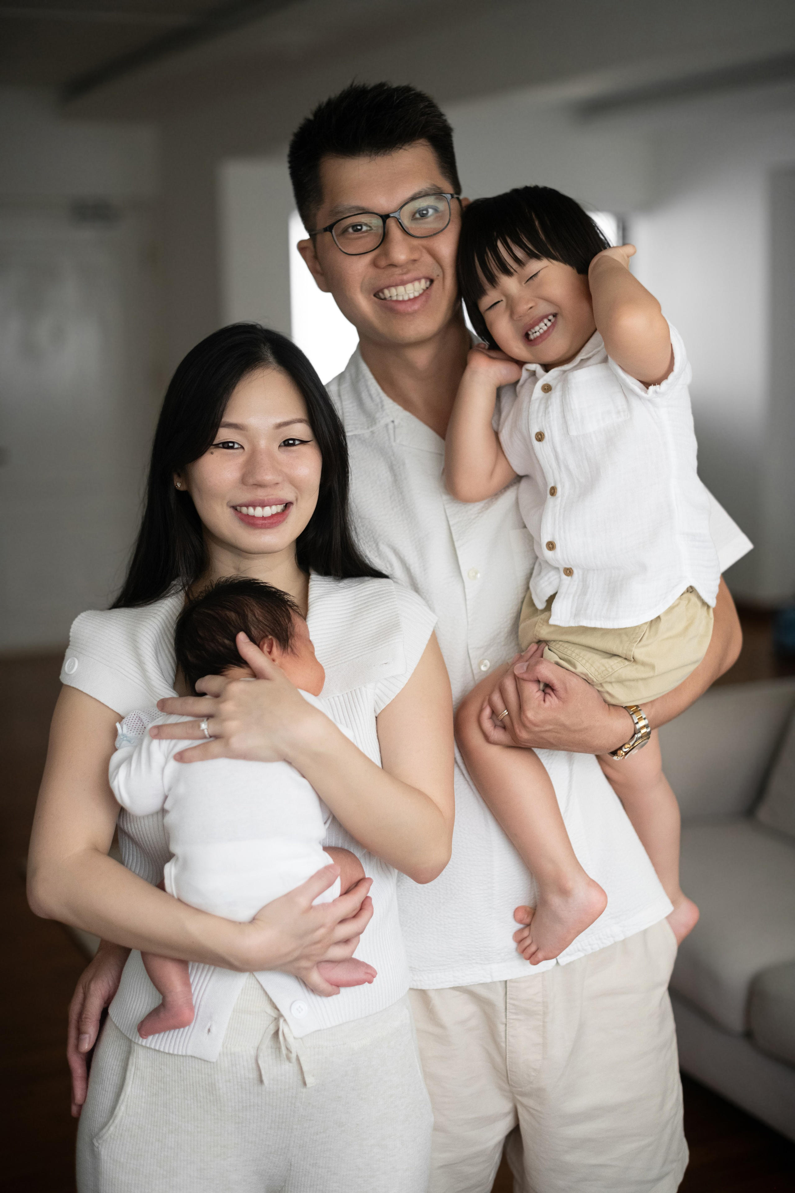 Lifestyle family portrait: parents holding newborn and laughing toddler, all in light neutrals indoors.
