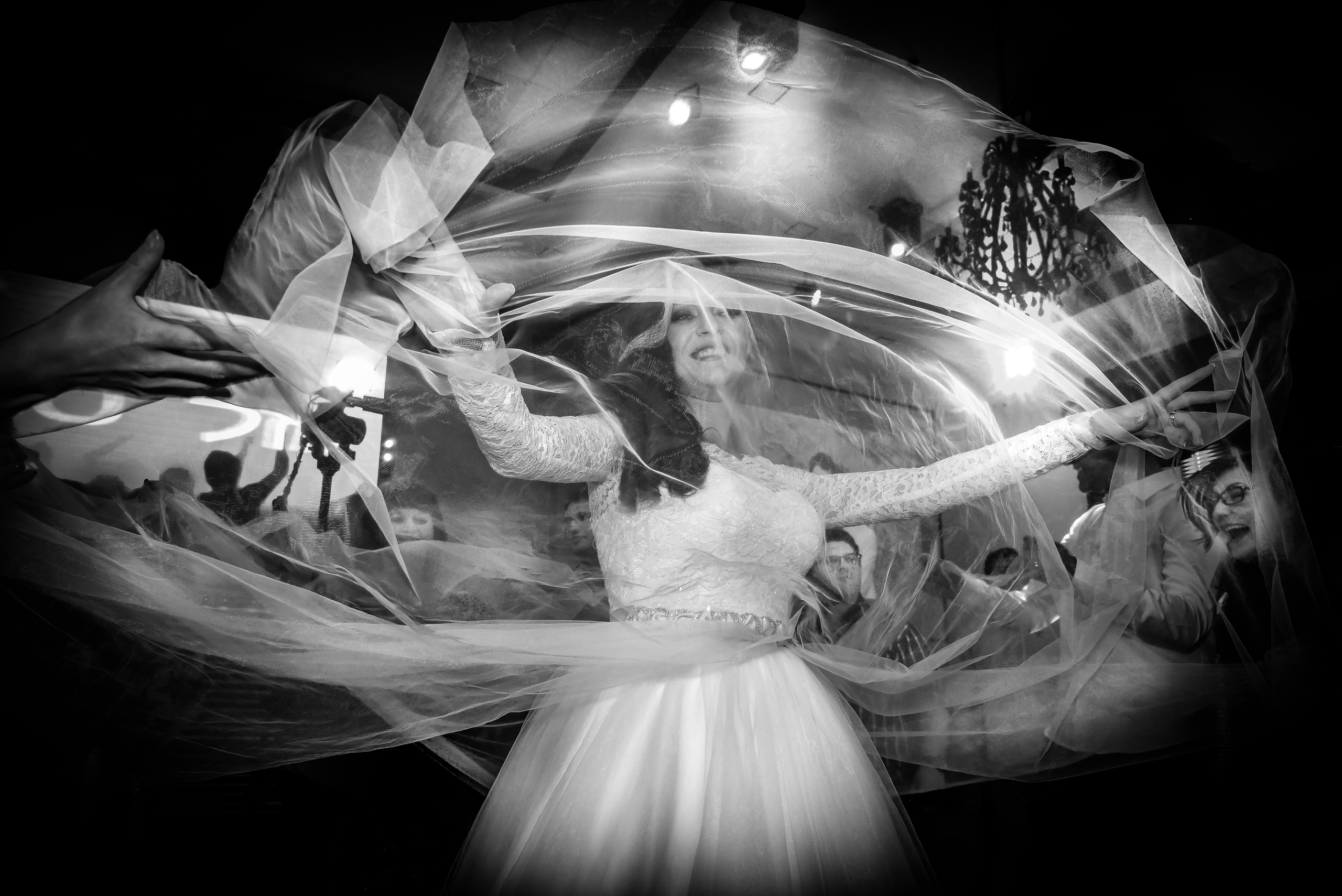 An emotional black-and-white shot: the bride at the center, wrapped in the movement of her veil. Her joy and the guests' reactions create a powerful celebratory atmosphere.