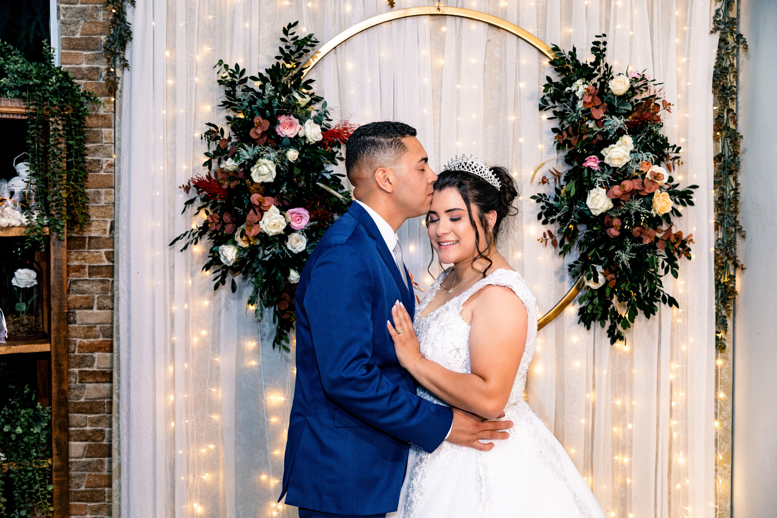Noivo beijando a testa da noiva em frente a um painel decorado com flores e luzes, cena íntima e simbólica.