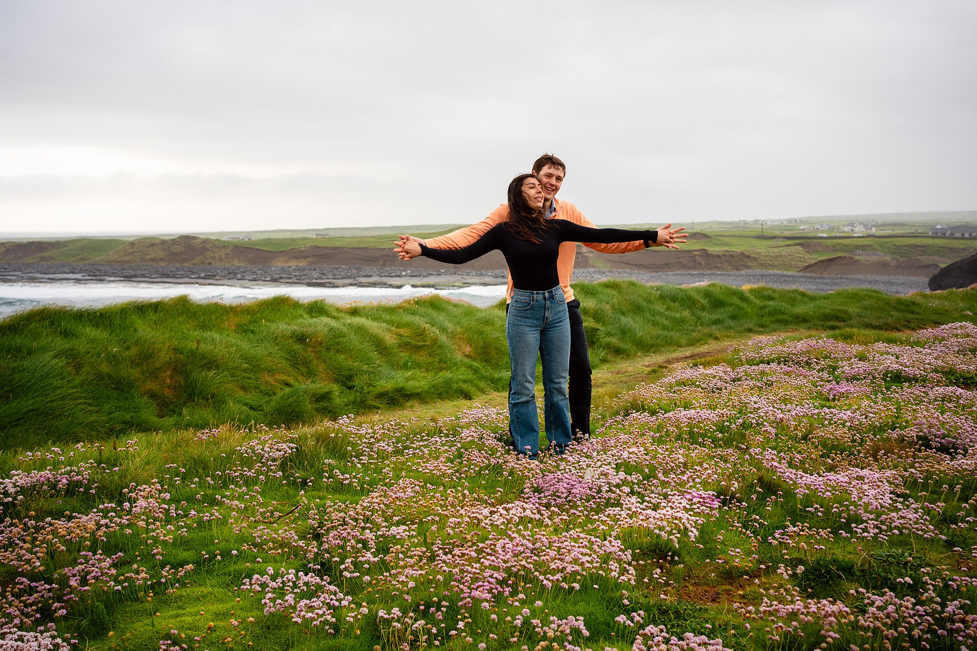A wide-angle view of a couple standing with their arms outstretched, enjoying the scenic wildflower fields of the Irish coast near Dublin.
