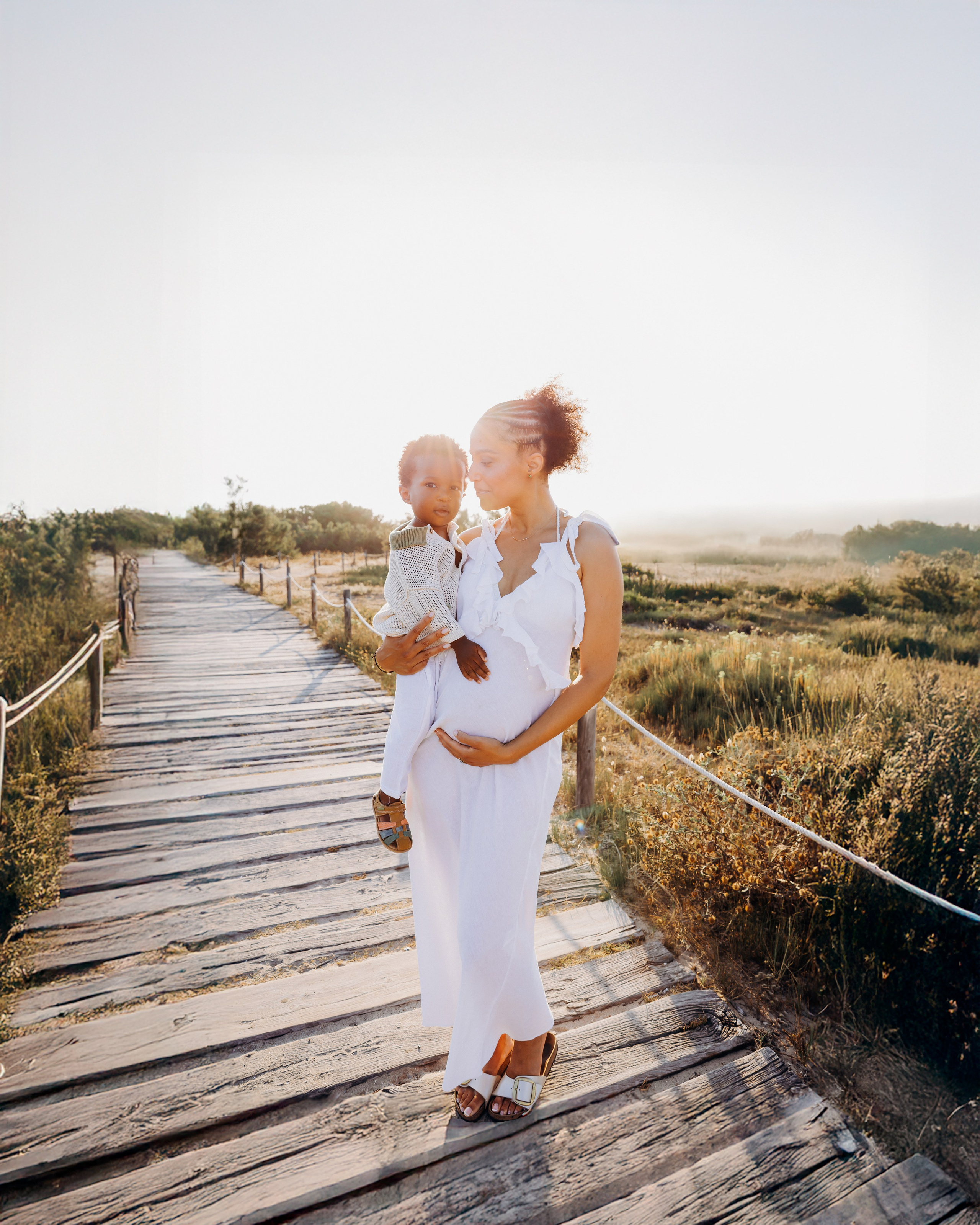 Madre embarazada sosteniendo a su hijo pequeño en una pasarela de madera al atardecer en Alicante, España — momento íntimo ideal para sesiones de maternidad y fotografía familiar en la costa alicantina.