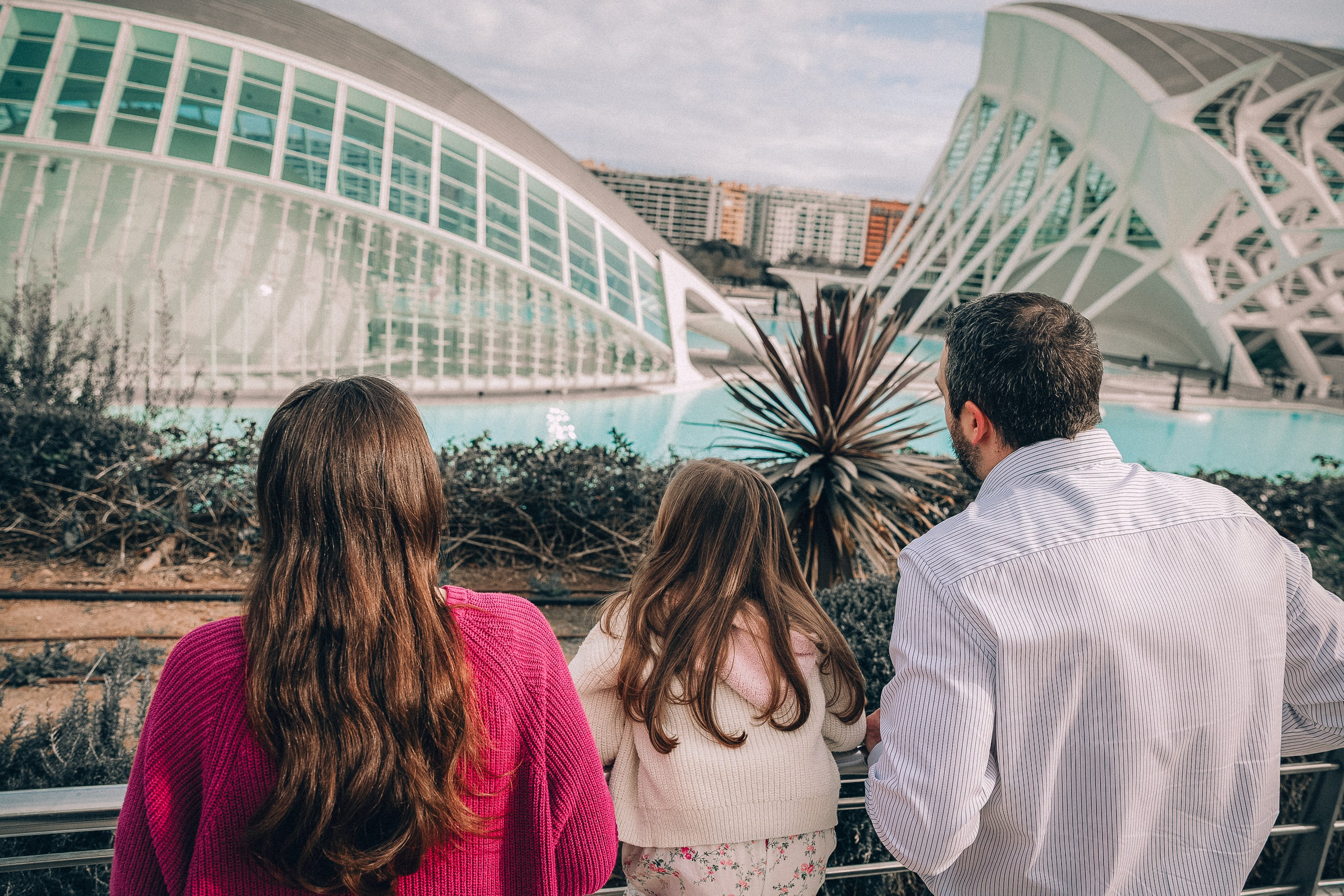 Семейная фотосессия в Валенсии, Испания, где родители и их ребёнок стоят вместе в Городе искусств и наук, снято со спины с культовой современной архитектурой, создавая lifestyle- и travel-настроение — идеально для семейных фотосессий в Валенсии и по всей Испании.