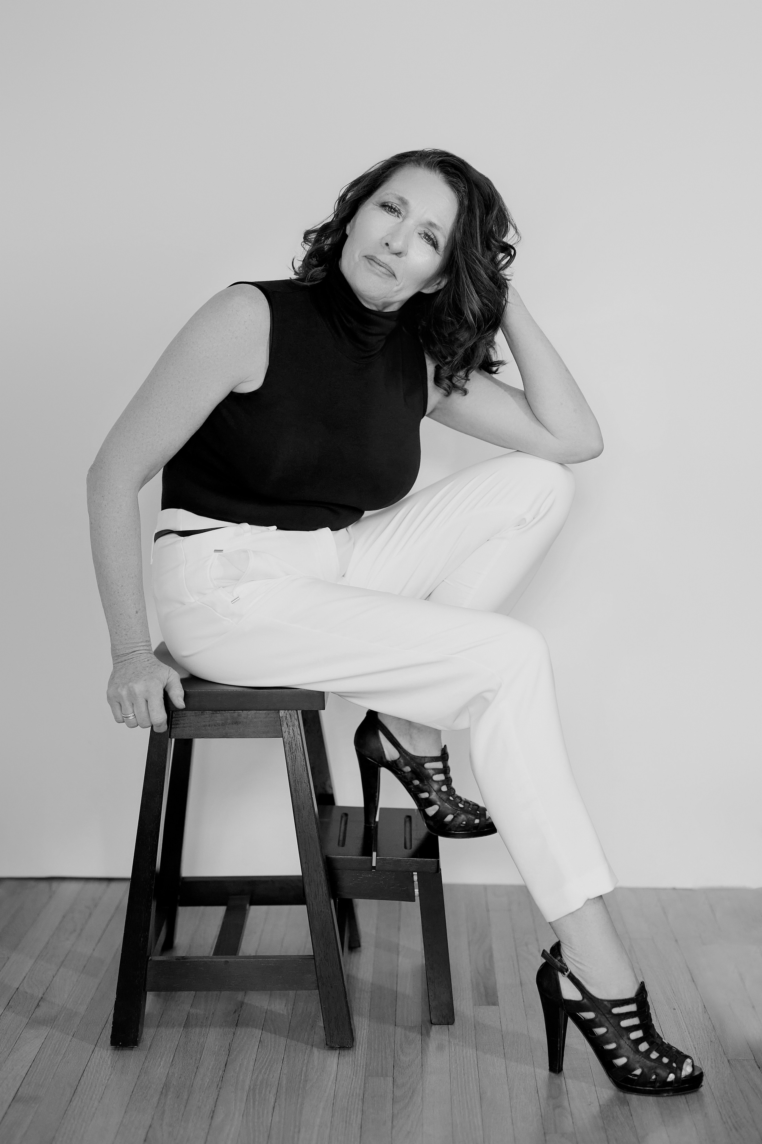 headshot for mature woman sitting on stool with white pants