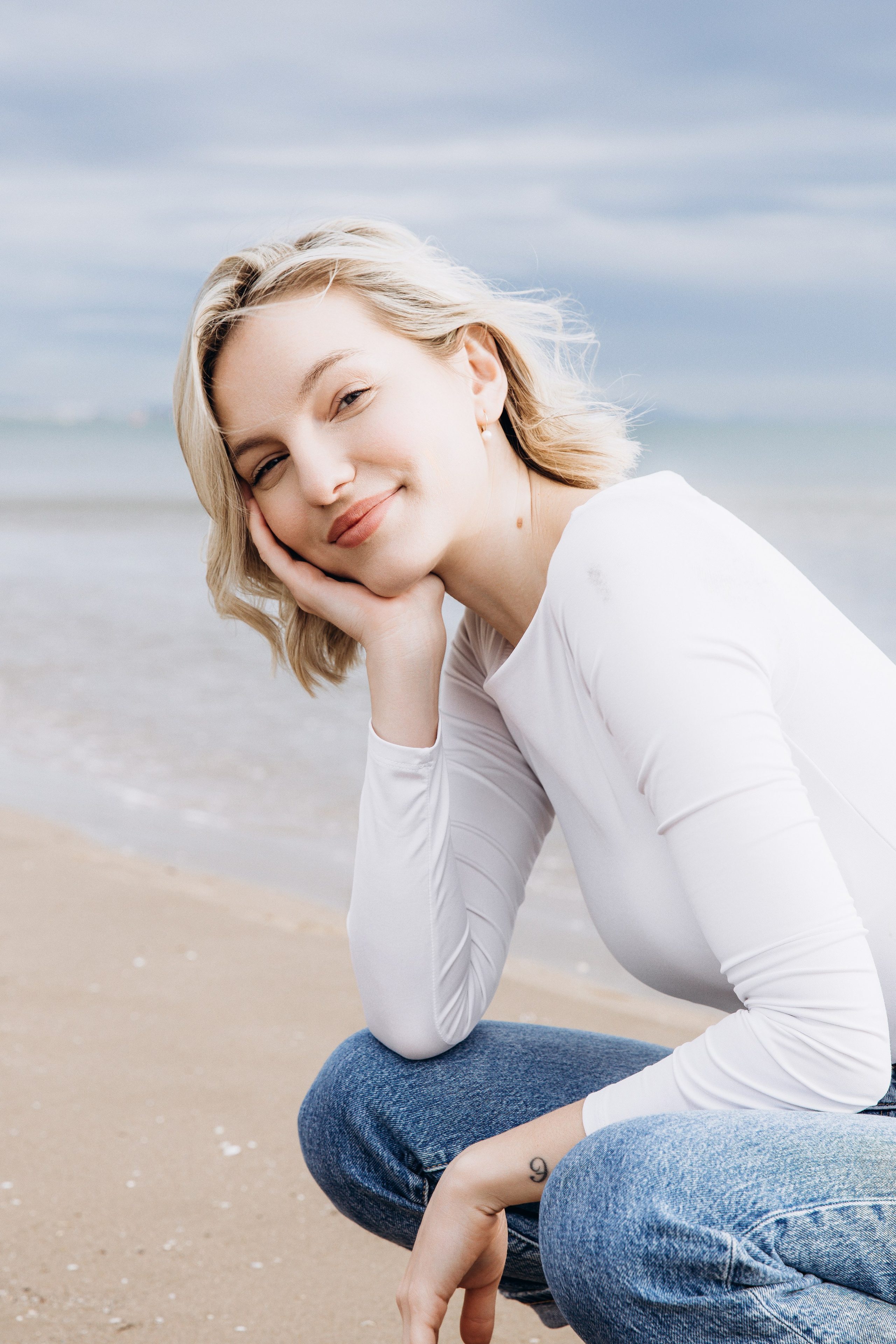 Непринуждённый женский портрет на пляже, снятый в Барселоне, Испания, с изображением блондинки в белом топе и джинсах, позирующей с нежной улыбкой — идеально подойдёт тем, кто ищет естественную и профессиональную женскую портретную фотосъёмку в Барселоне и Испании.