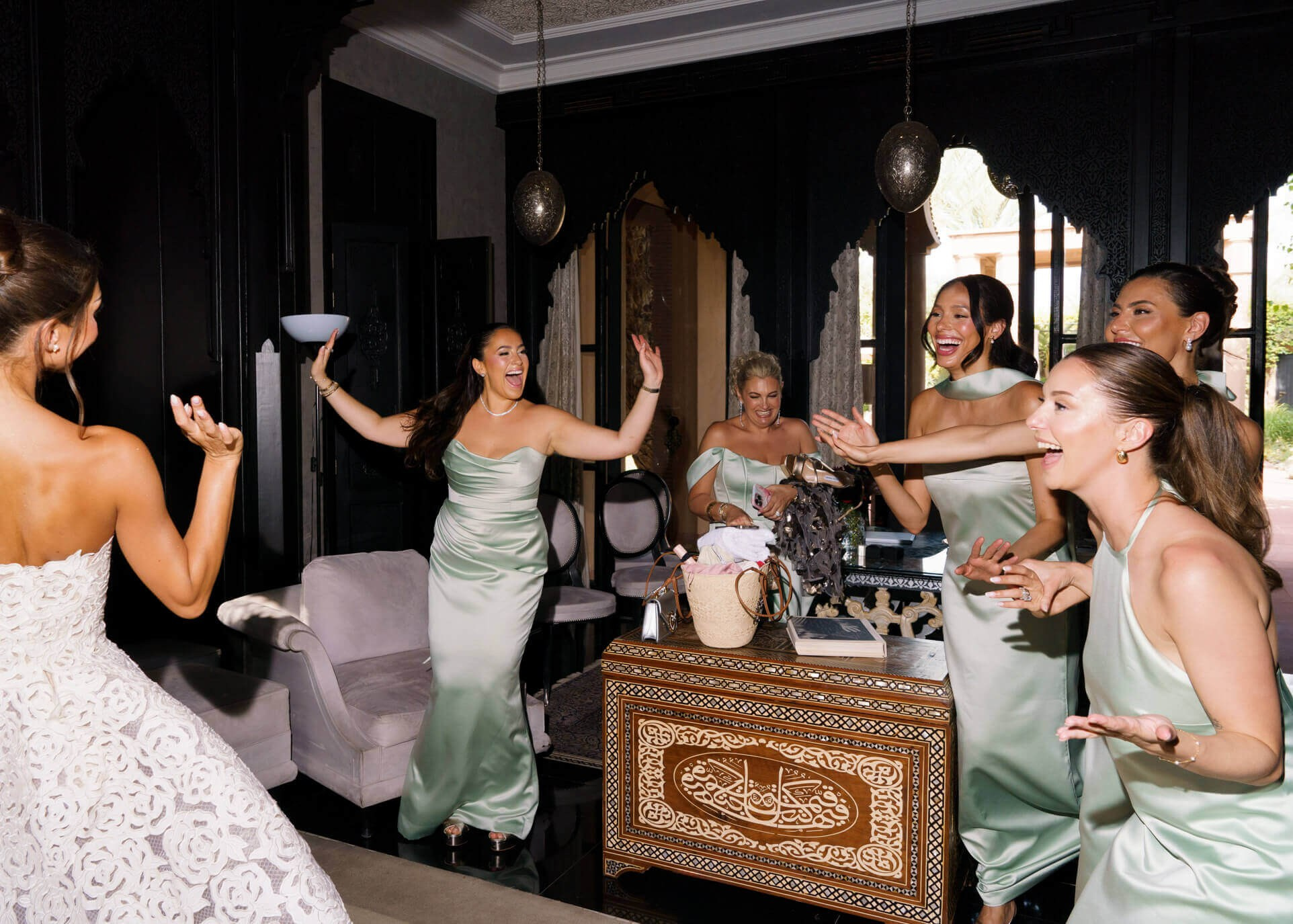 Bride in a white lace gown dancing with bridesmaids in sage green satin inside a Moorish riad salon, joyful getting-ready celebration in Morocco