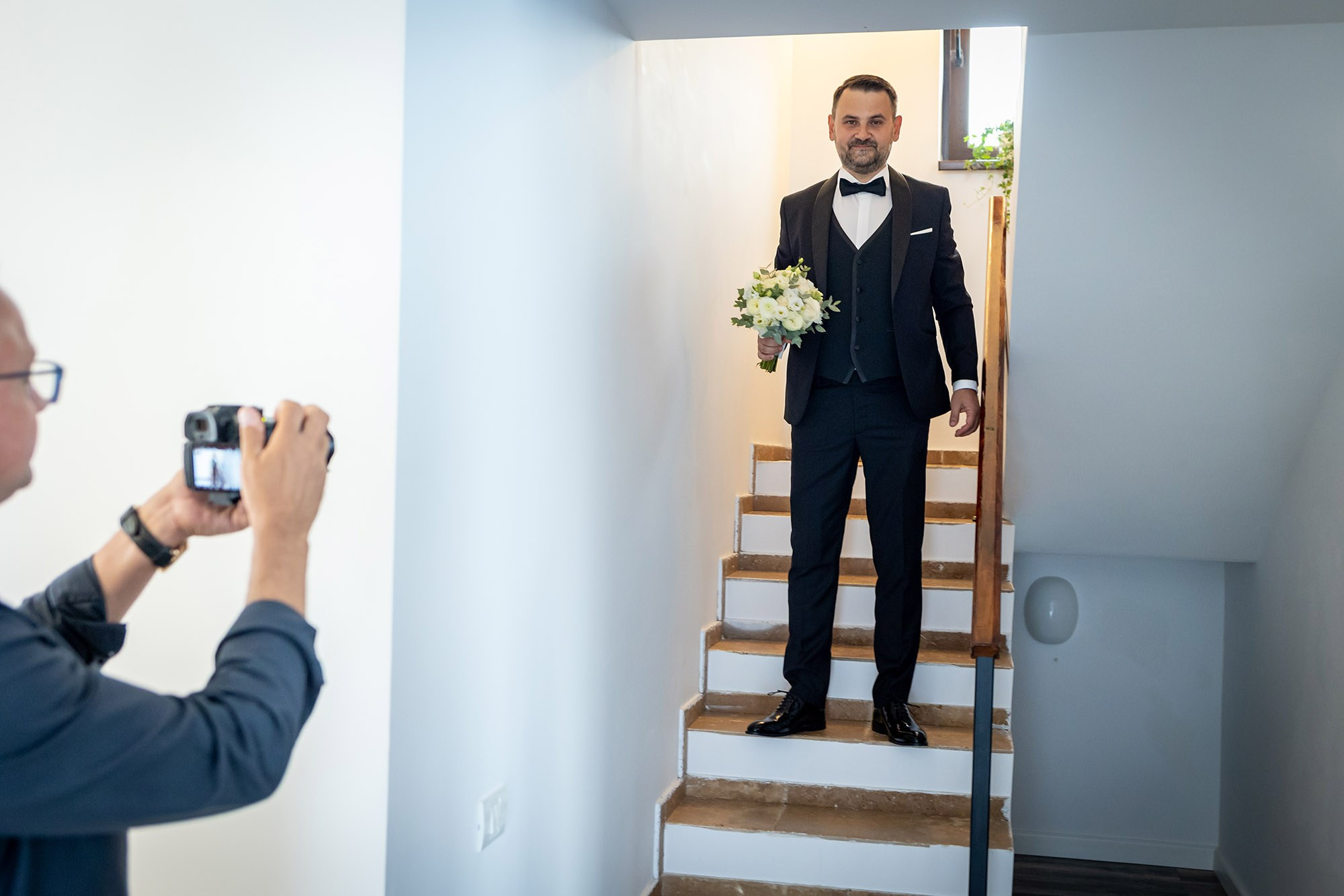 Mire elegant în costum negru cu papion, ținând buchetul de flori albe pe scară înainte de nuntă