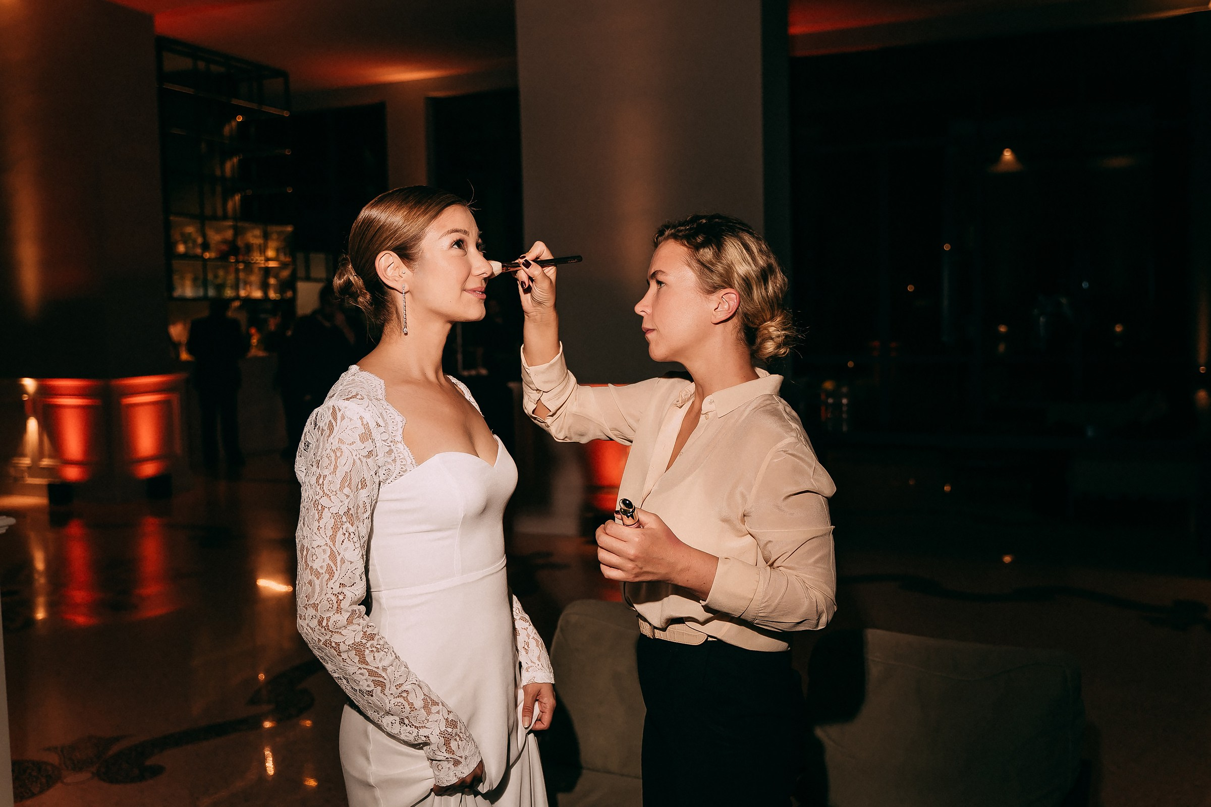 Bride receiving a makeup touch-up, standing poised in a white dress with lace sleeves, in an elegantly lit indoor venue.