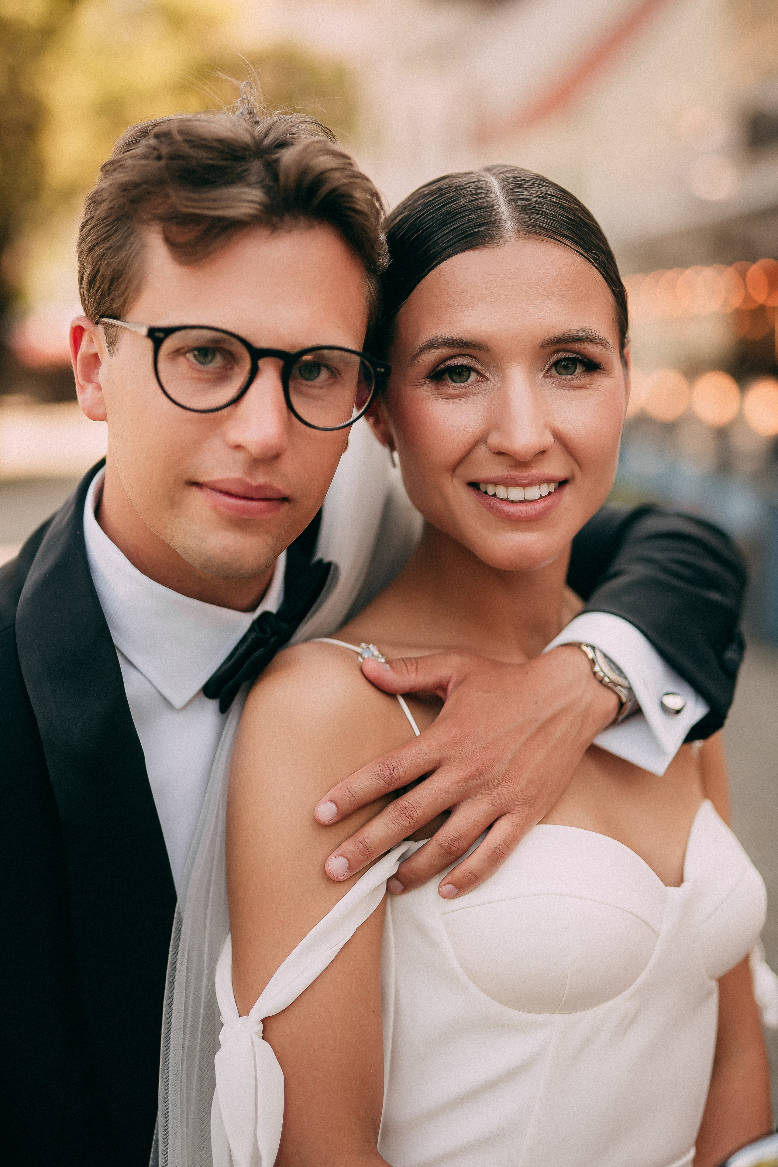 A couple in formal attire, with the man in a black tuxedo and the woman in a white off-shoulder dress, pose closely together. The background features blurred lights and greenery, suggesting an outdoor setting at a special event.