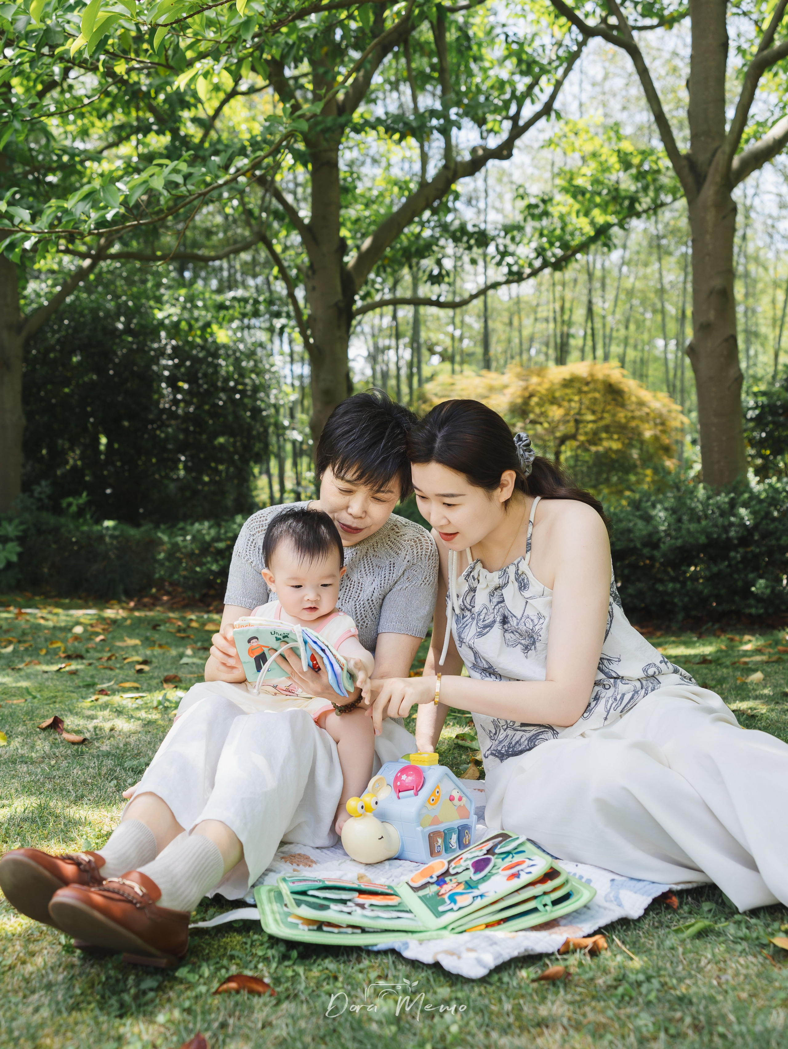 A mother and grandmother sit with a baby on a picnic blanket beneath green trees in a Shanghai park.
