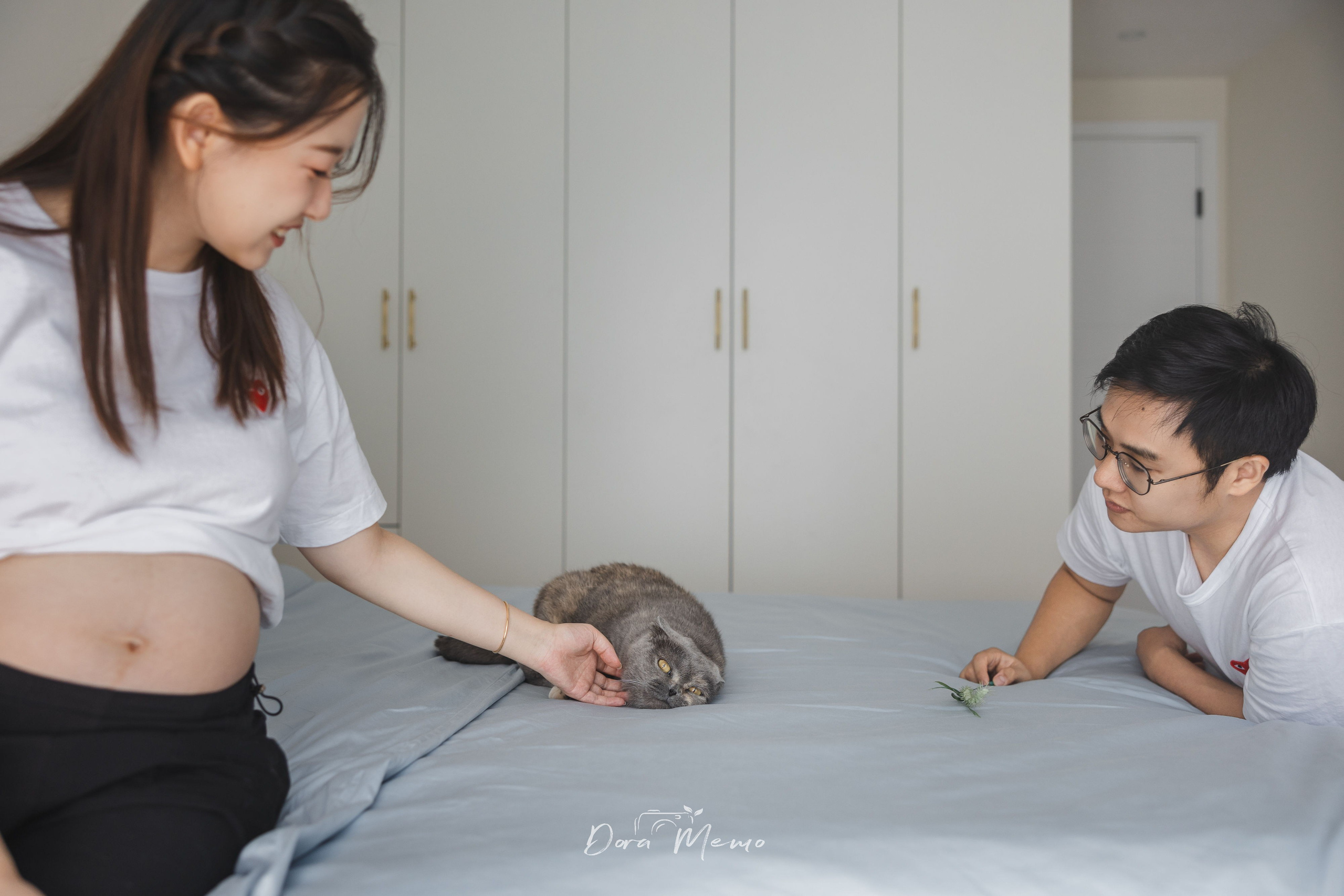 Expecting parents lie beside their cat on the bed in a calm lifestyle maternity photograph.