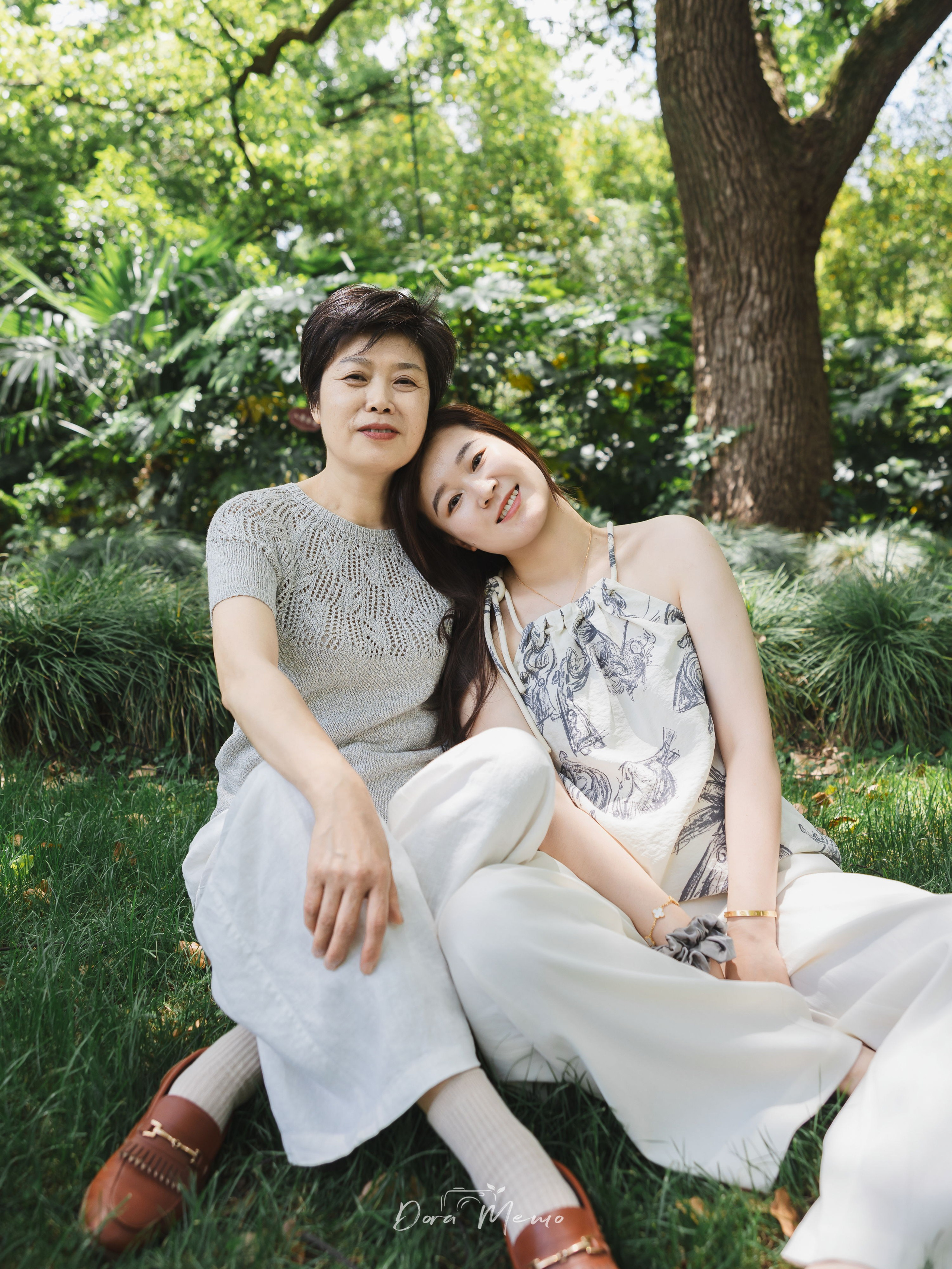 A mother rests her head on her own mother’s shoulder in a quiet portrait on the grass.