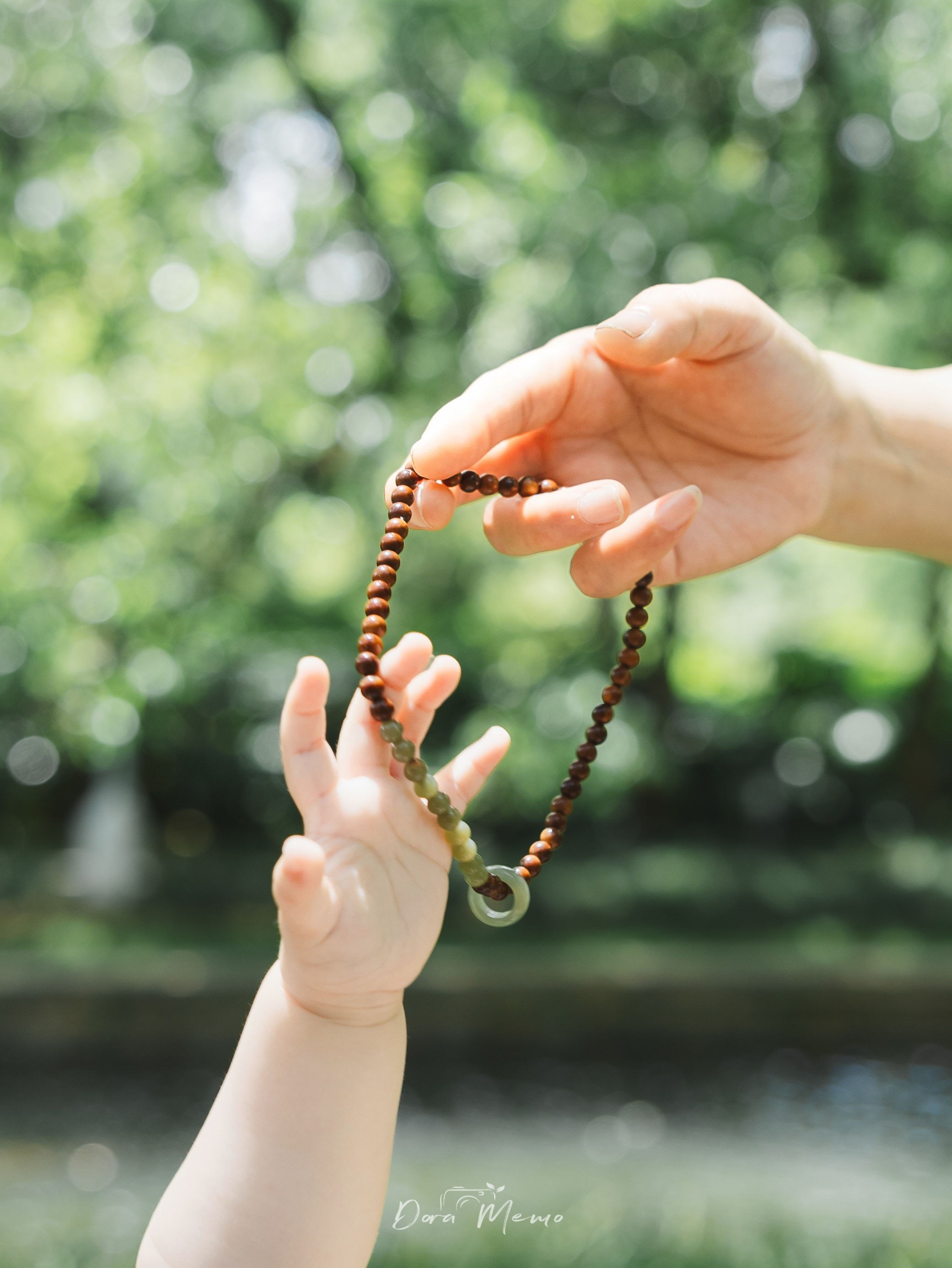A baby reaches toward a beaded bracelet held by an adult hand in bright natural light.