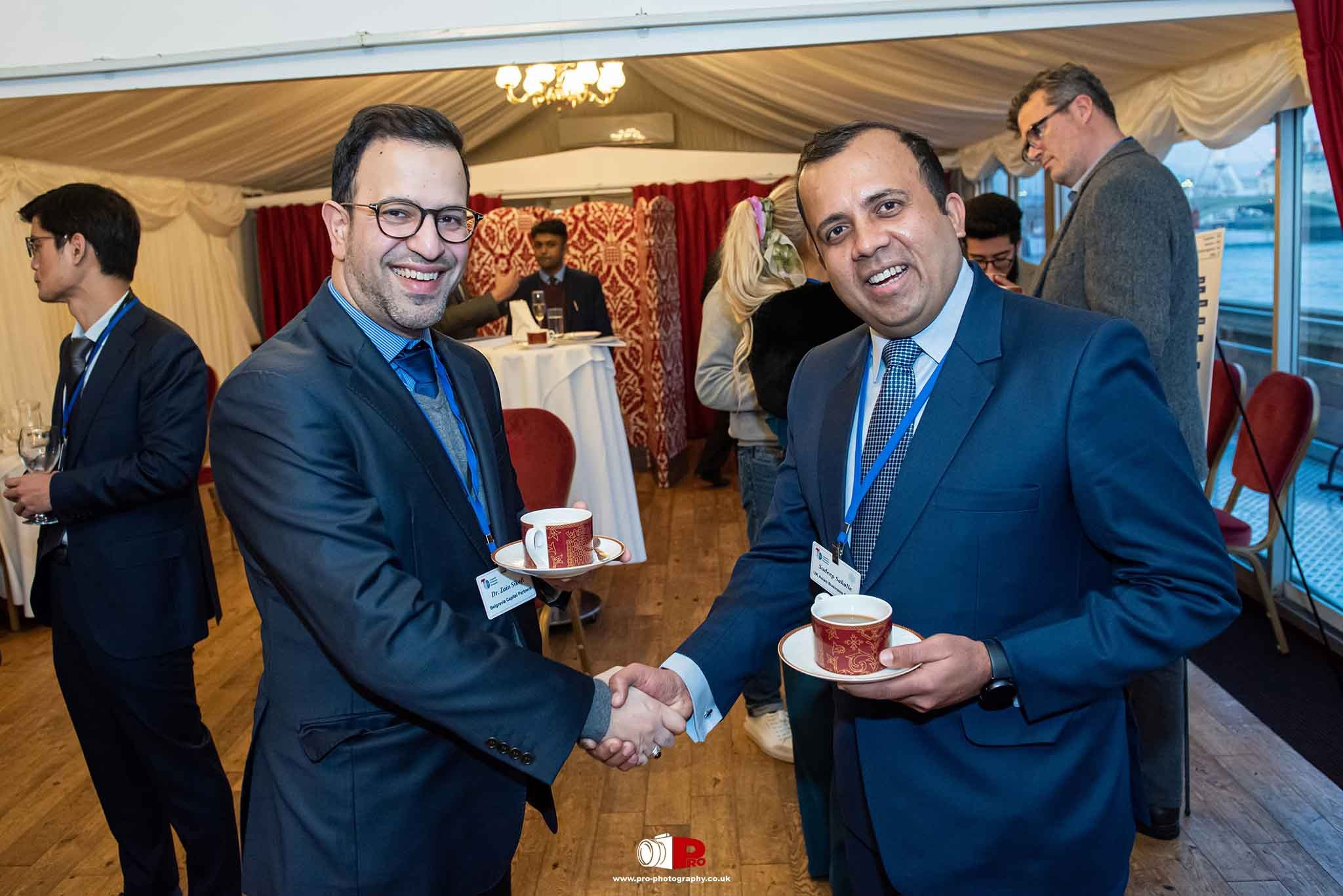Two men in suits smiling and shaking hands at a professional networking event.
