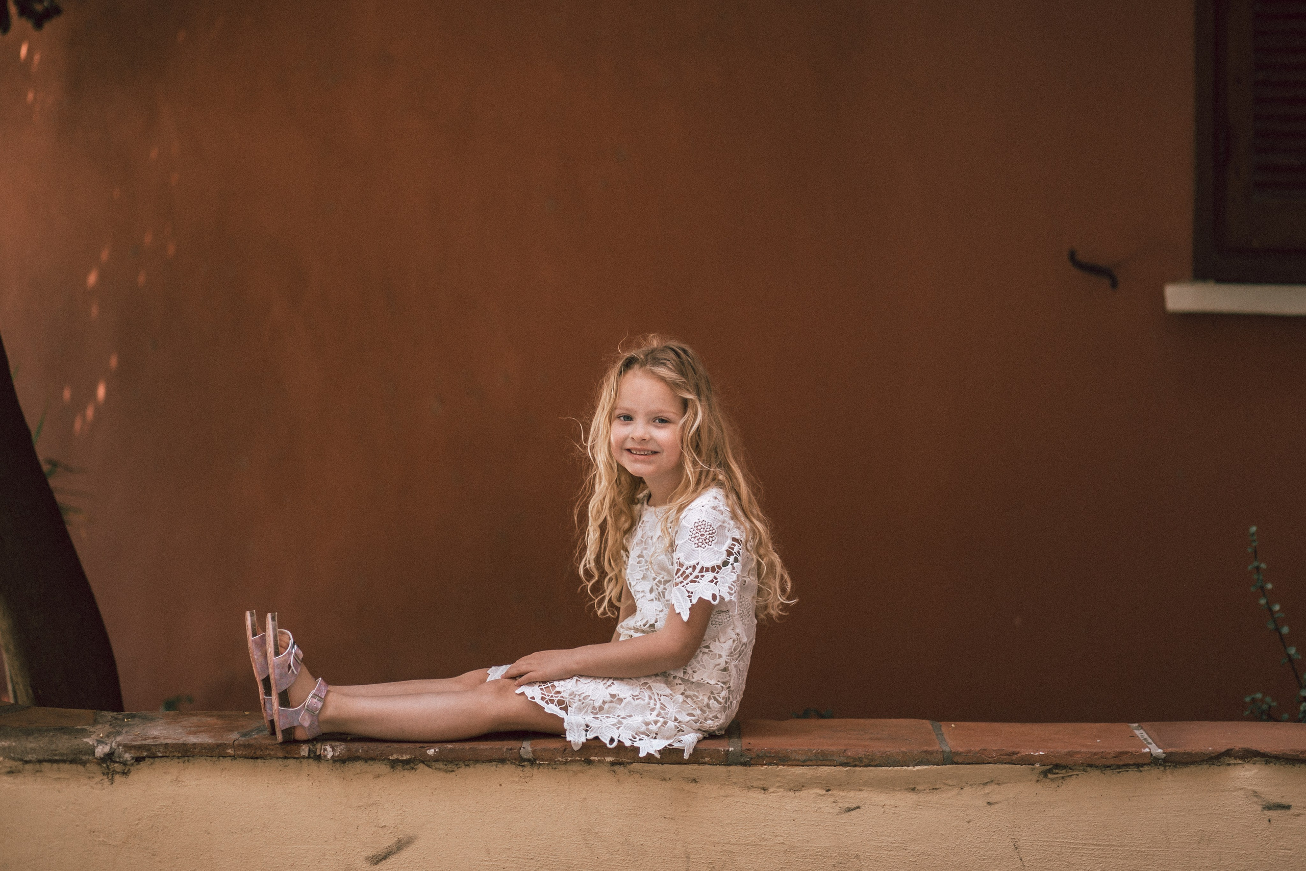 Портрет улыбающейся молодой девушки с длинными светлыми волосами, в белом кружевном платье и сандалиях, сидящей на низкой стене на фоне деревенского оранжевого фона в Валенсии, Испания